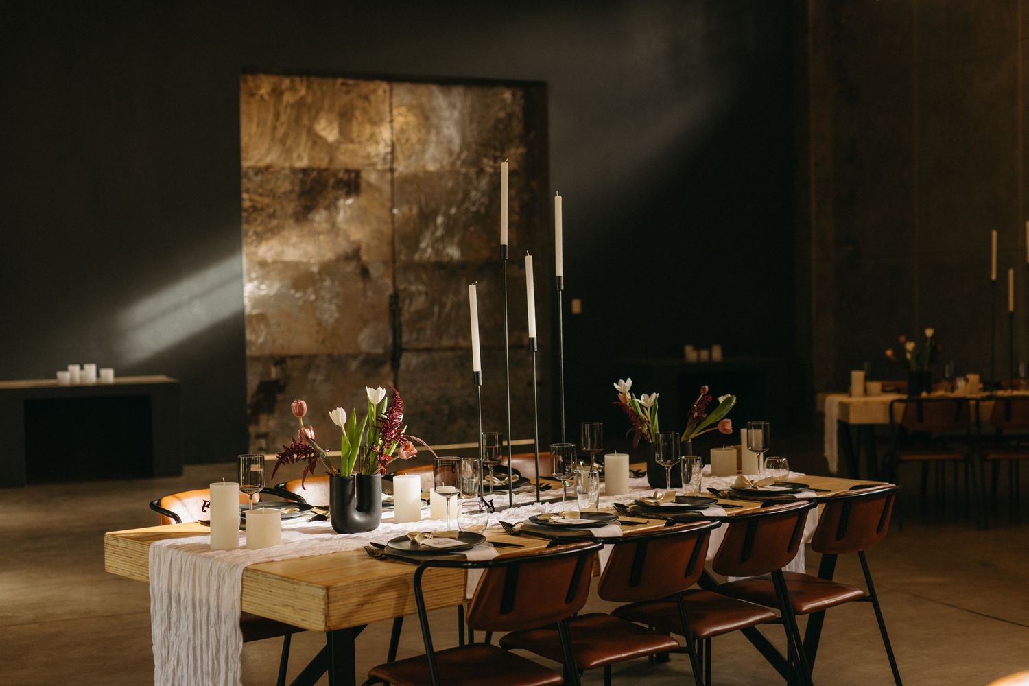 A rustic wedding reception table setup with candles and floral arrangements in a dark industrial venue with sunbeams streaming in.