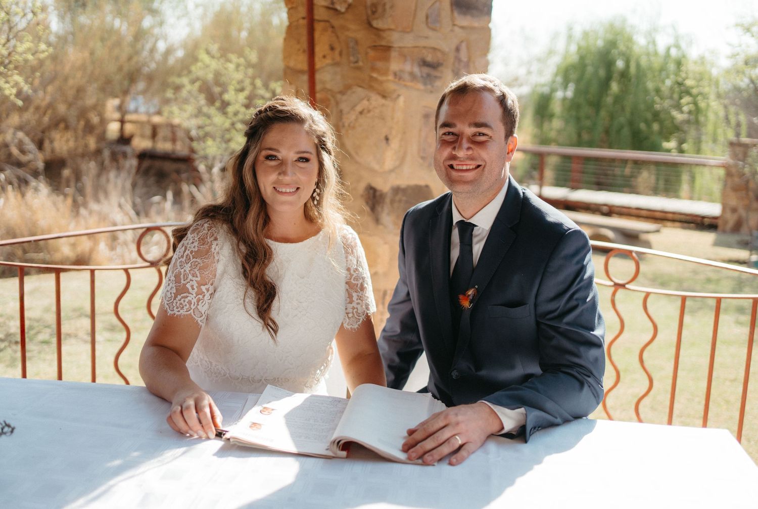 Newlyweds sitting at table signing marriage documents at outdoor venue.