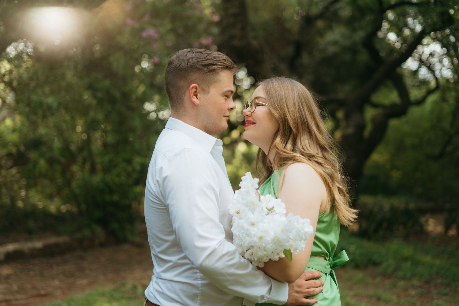 A romantic outdoor portrait of a couple embracing while holding white flowers in a garden setting.