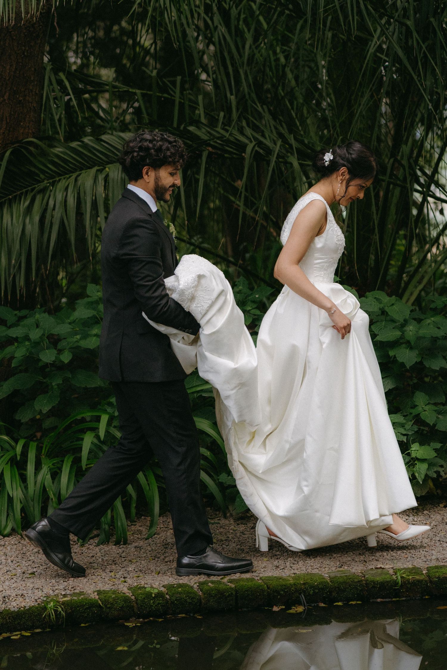 Wedding couple navigating a garden path together, with groom helping bride with her flowing white dress.