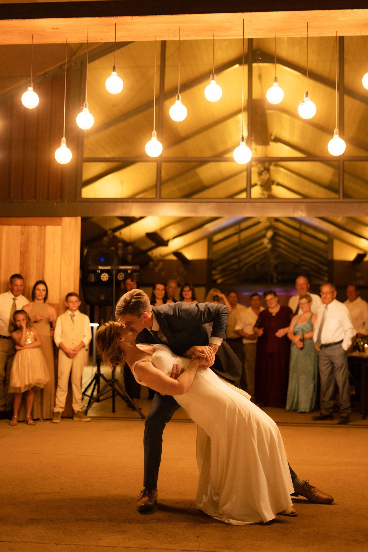 A couple performs their first dance under warm string lights in a rustic venue with wooden beams and onlooking guests.
