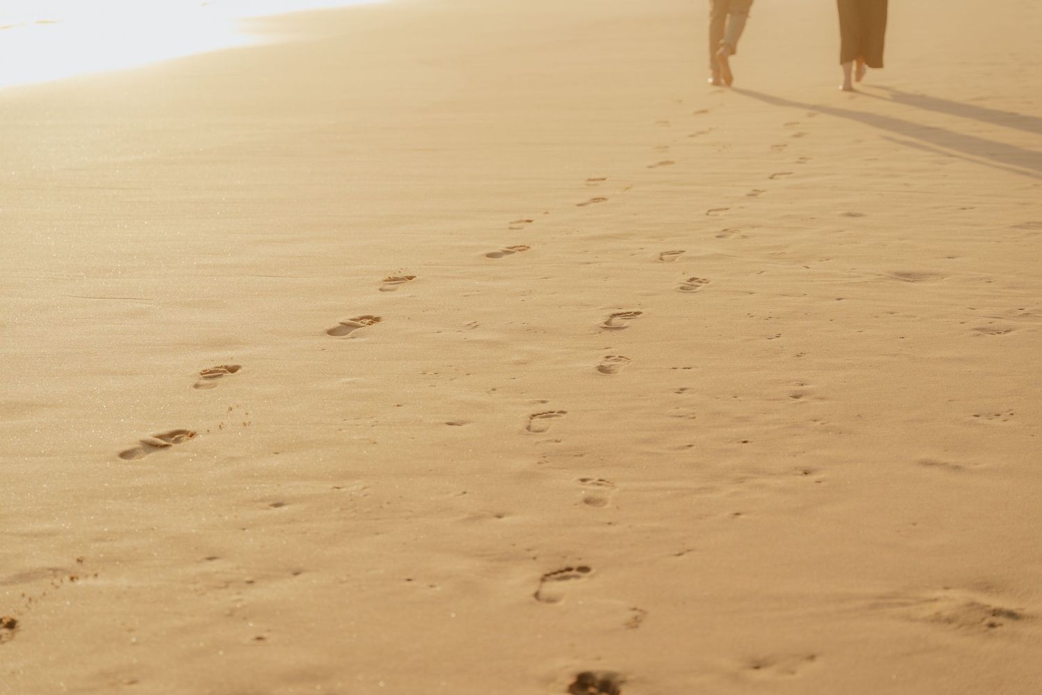 Footprints trail across a sandy beach at sunset with legs of a person walking visible in the warm golden light.