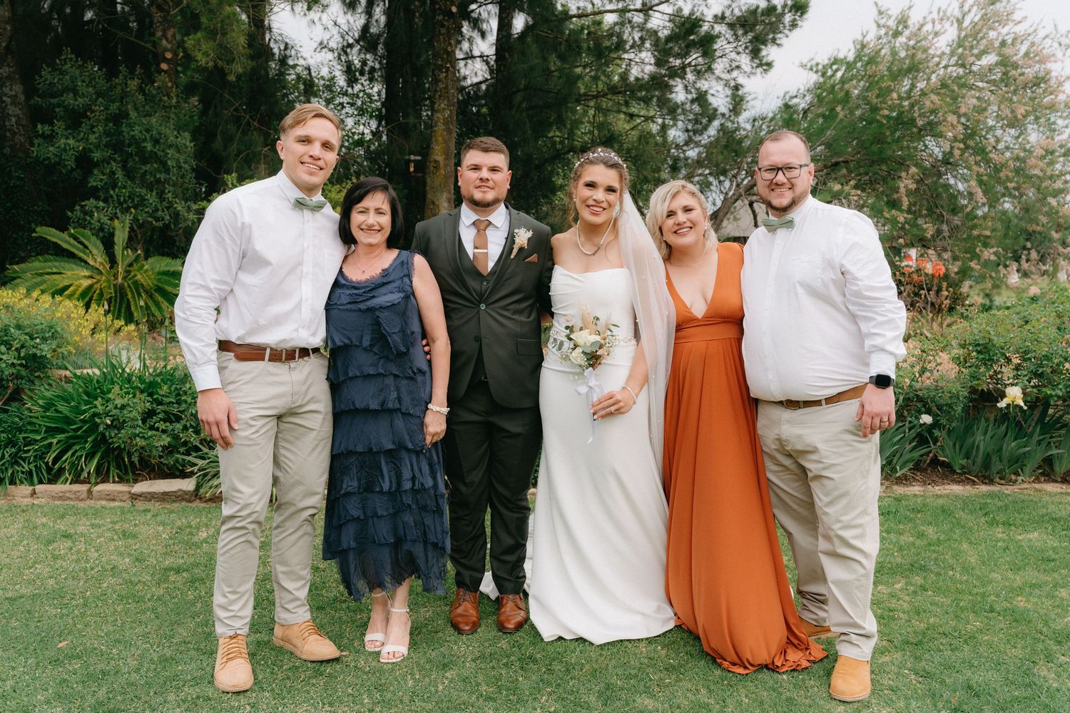 A group of people in coordinated wedding attire pose together on a grassy lawn during an outdoor celebration.