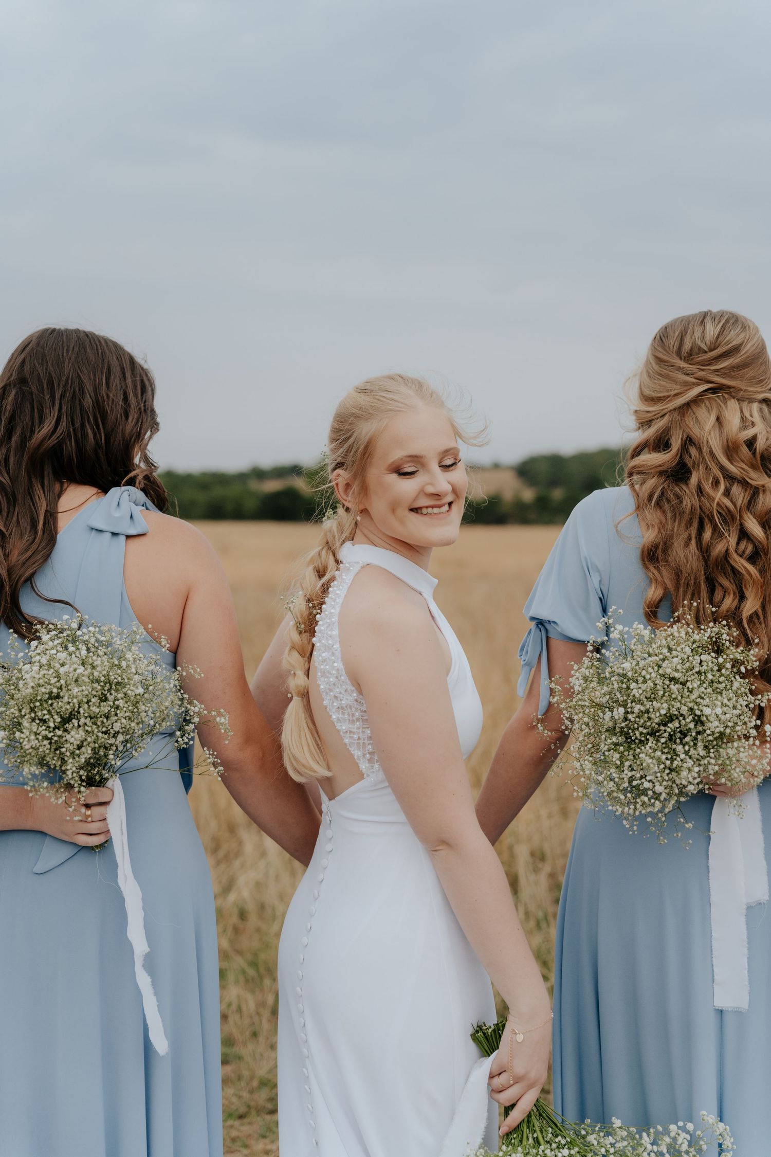 Bridesmaids in light blue dresses hold baby's breath bouquets against a rural field backdrop.