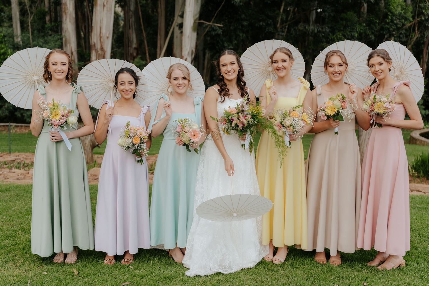 Bridesmaids in pastel colored dresses stand together holding floral bouquets during an outdoor wedding ceremony.