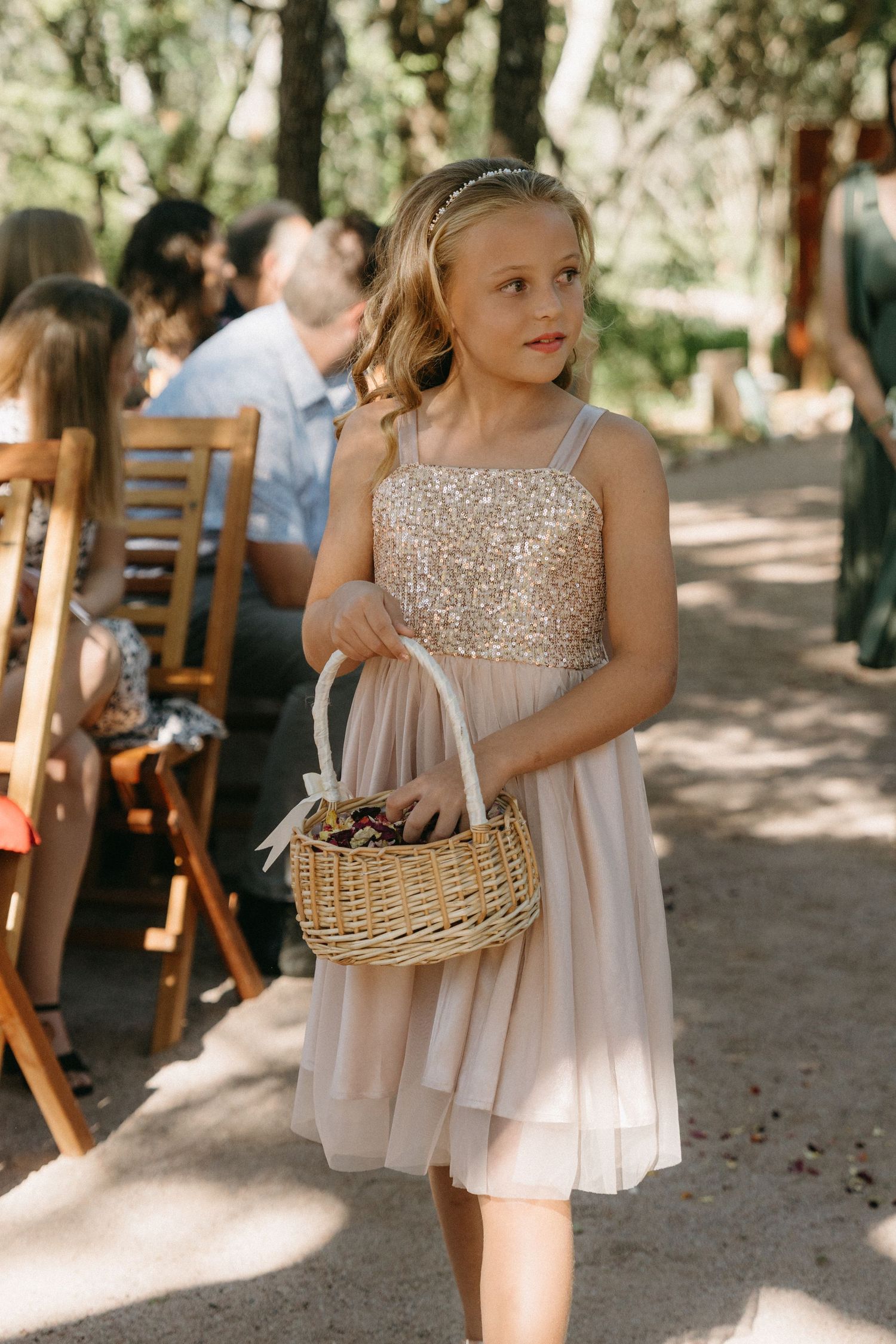 Girl in sparkly dress carries flower basket along outdoor ceremony aisle.