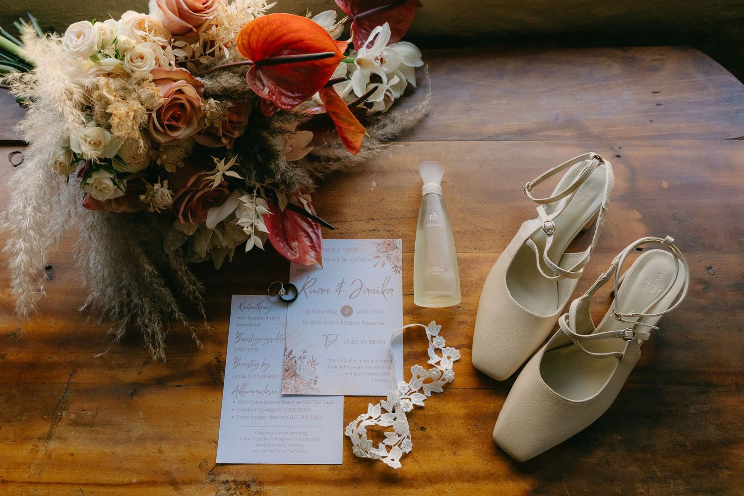 Wedding details flat lay featuring invitation, shoes, and floral elements on wooden surface.