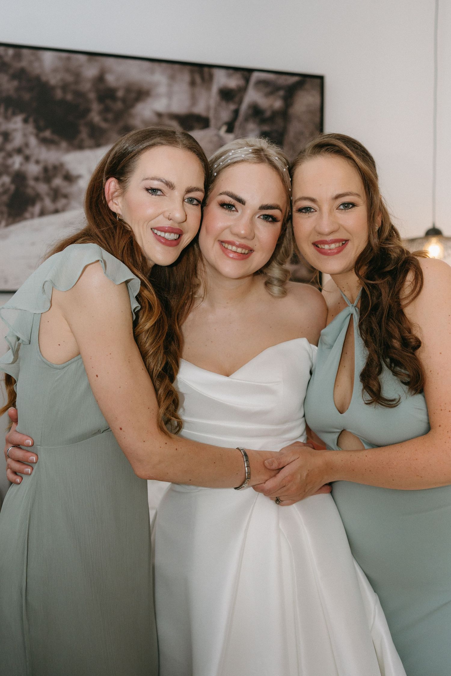 Three women pose together in pastel mint bridesmaid dresses and a white wedding gown at an indoor celebration.