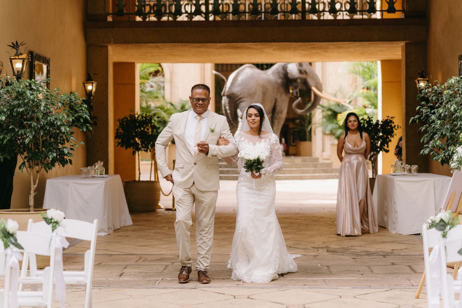 Wedding ceremony setup at elegant venue with white chairs, floral arrangements and elephant statue visible in background.