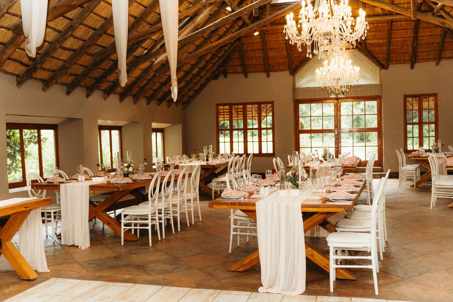 Elegant rustic wedding reception venue with crystal chandeliers, wooden beams, and white draped tables set for dining.