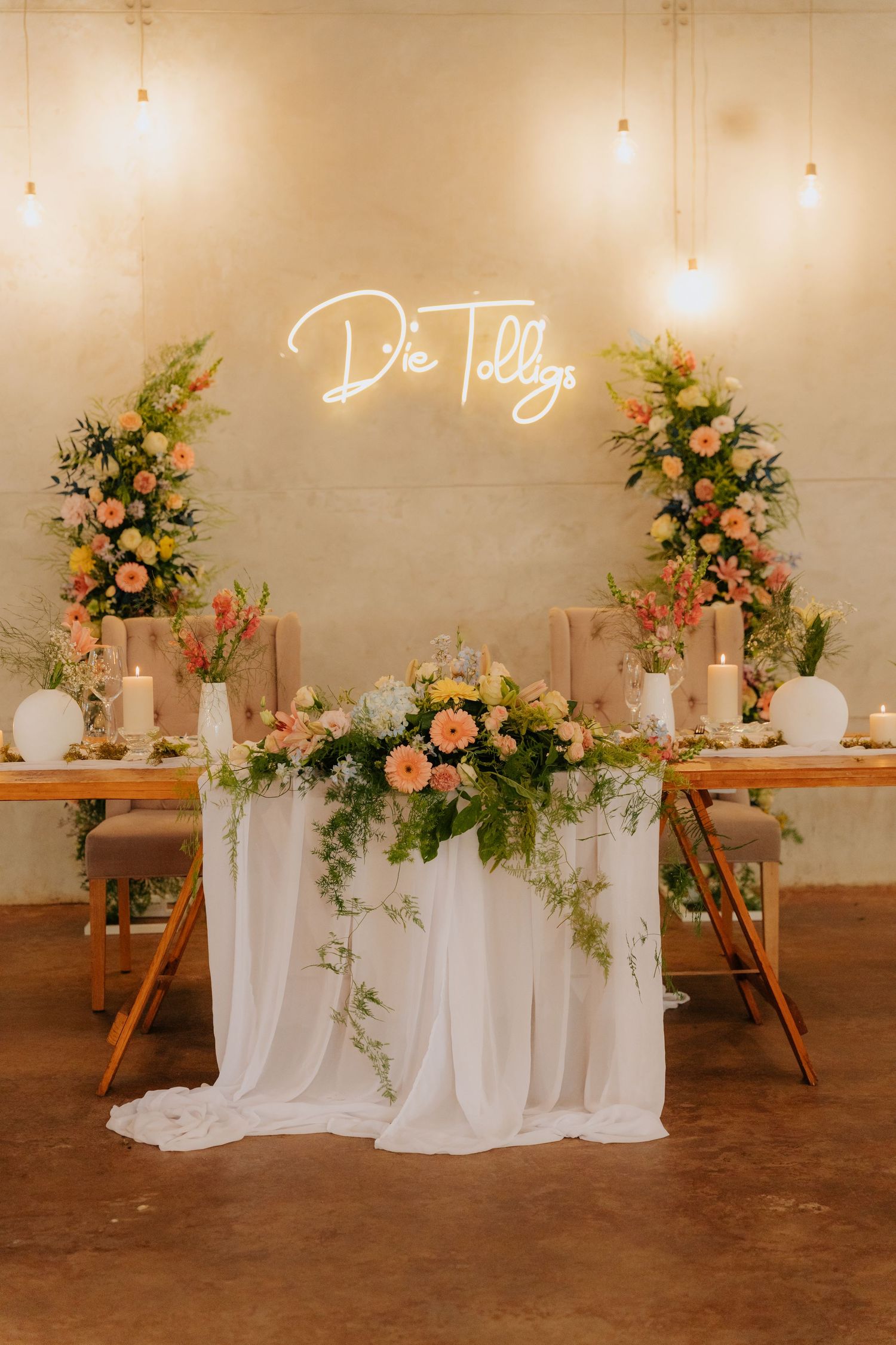 Elegant wedding reception @Boshoek, Mpumalanga with white draping, peach and coral floral arrangements, and neon 'Die Tolligs' sign above.