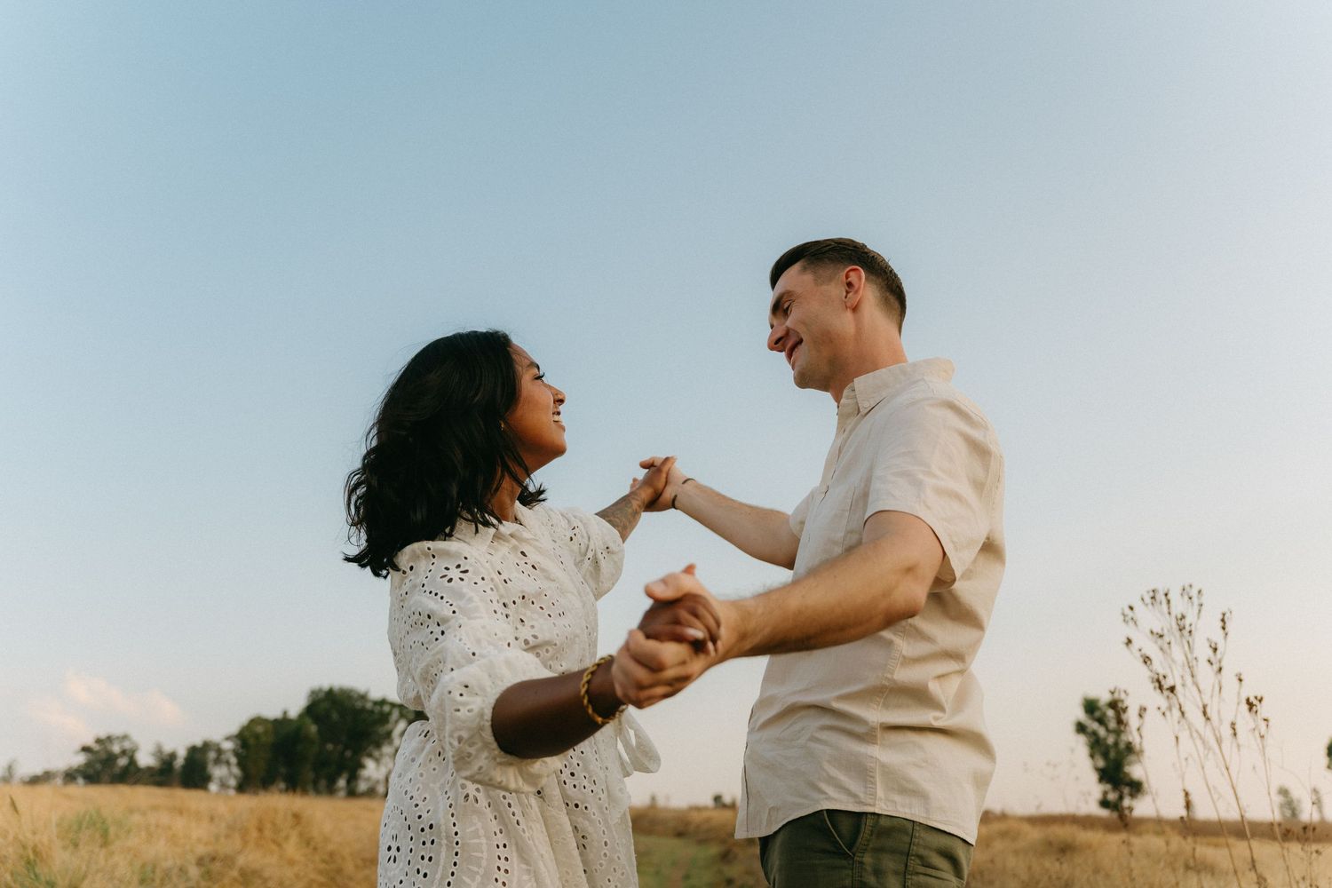 A couple in white outfits dancing together joyfully in a sunlit outdoor setting at golden hour.