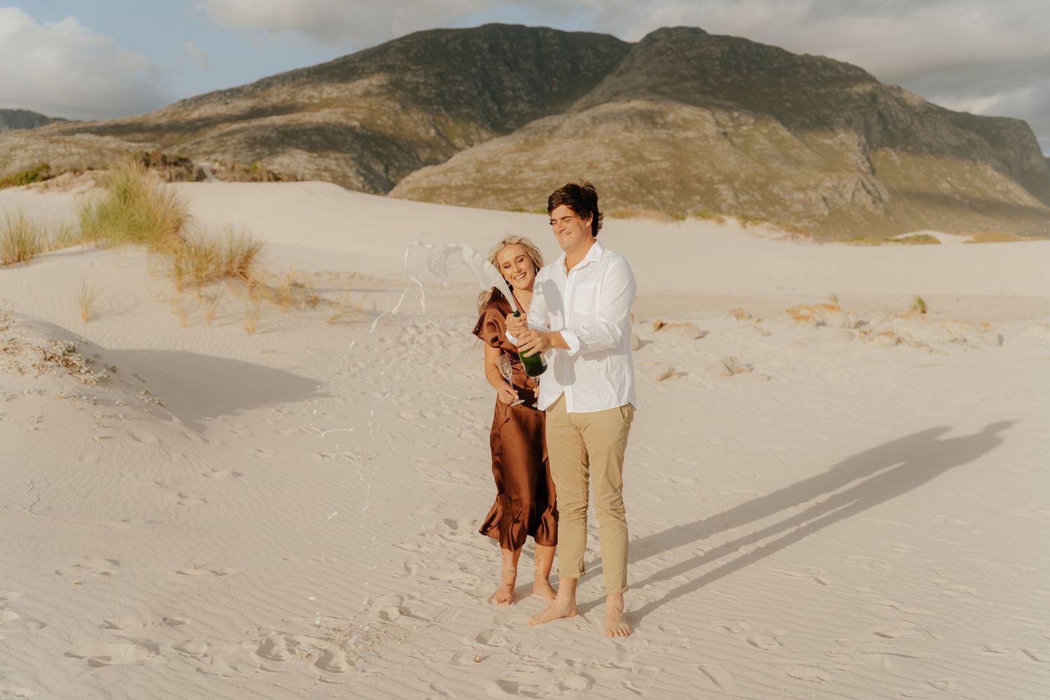 Couple embracing on white sand beach with mountain backdrop, woman in brown dress, man in white shirt.
