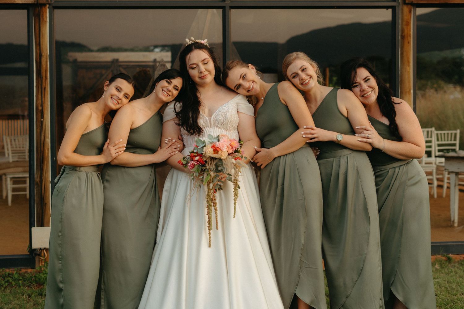 Wedding party posing together in sage green bridesmaid dresses with bride holding autumn-colored bouquet.