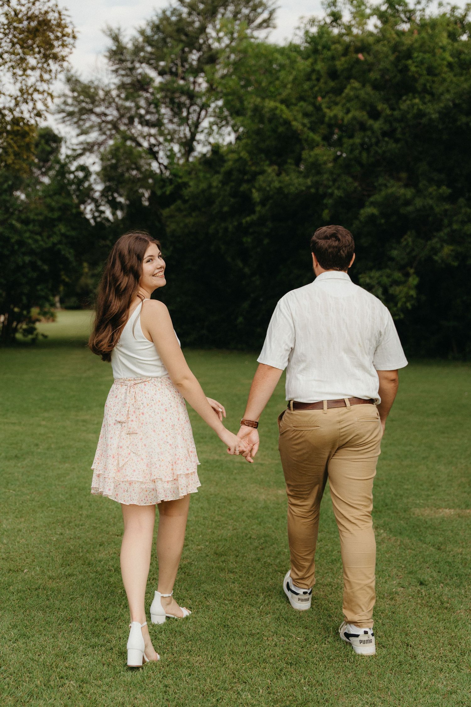 A smiling young woman in a floral skirt holds hands with a man as they walk through a lush green park.