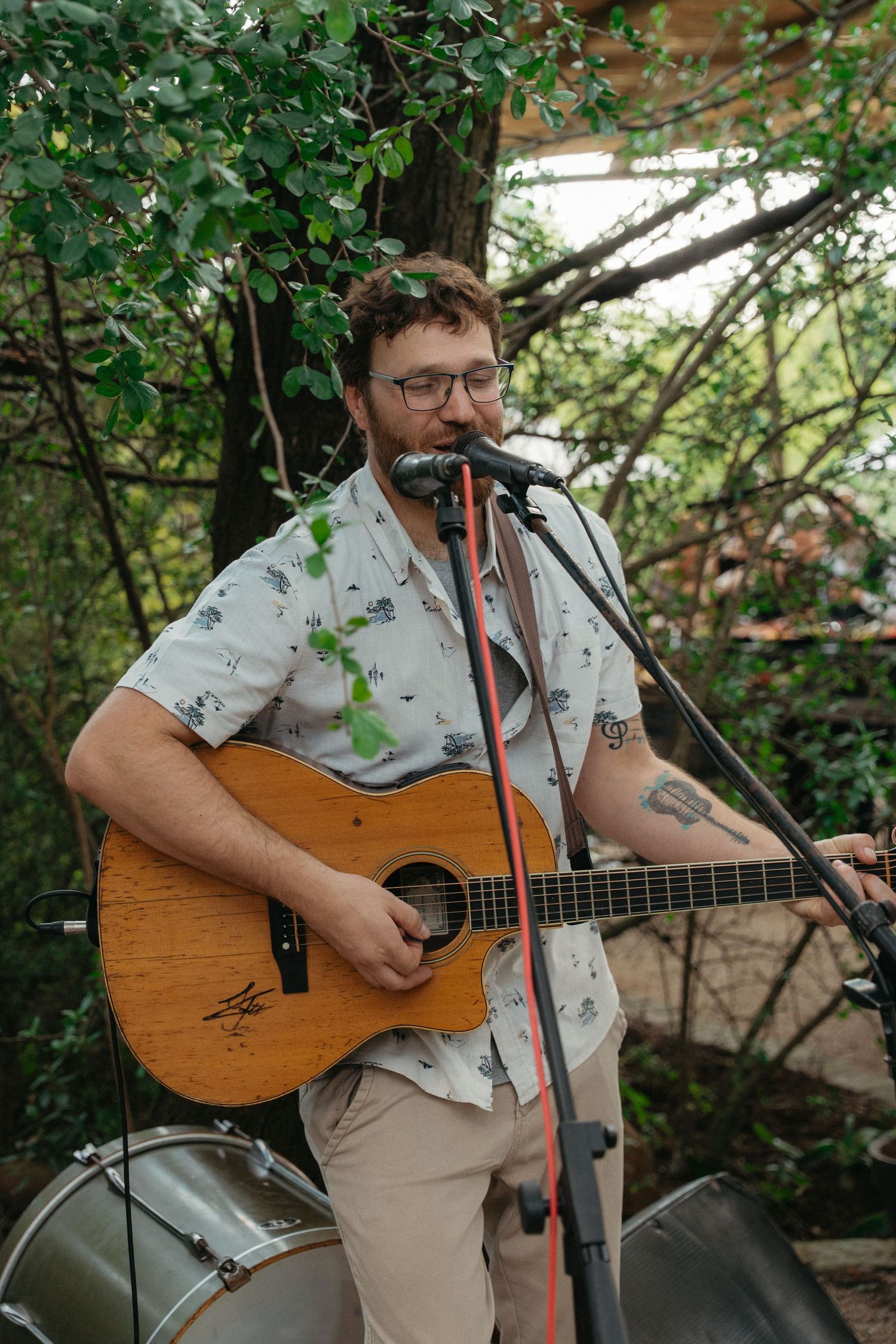A musician performs outdoors with an acoustic guitar wearing a floral print shirt and khaki pants.