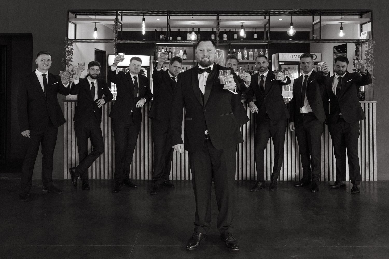 A vintage black and white photograph shows a group of men in formal attire standing at an elegant bar counter.