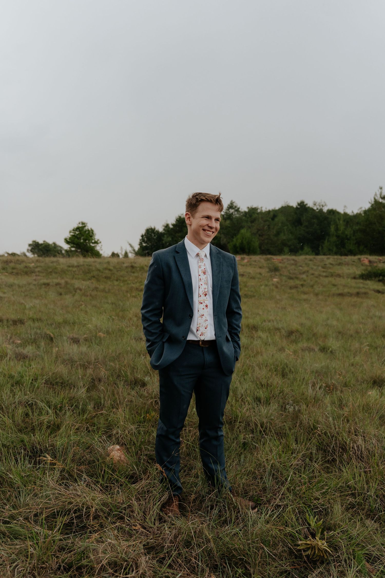 A person wearing a navy suit stands in a rustic grassy field on an overcast day.