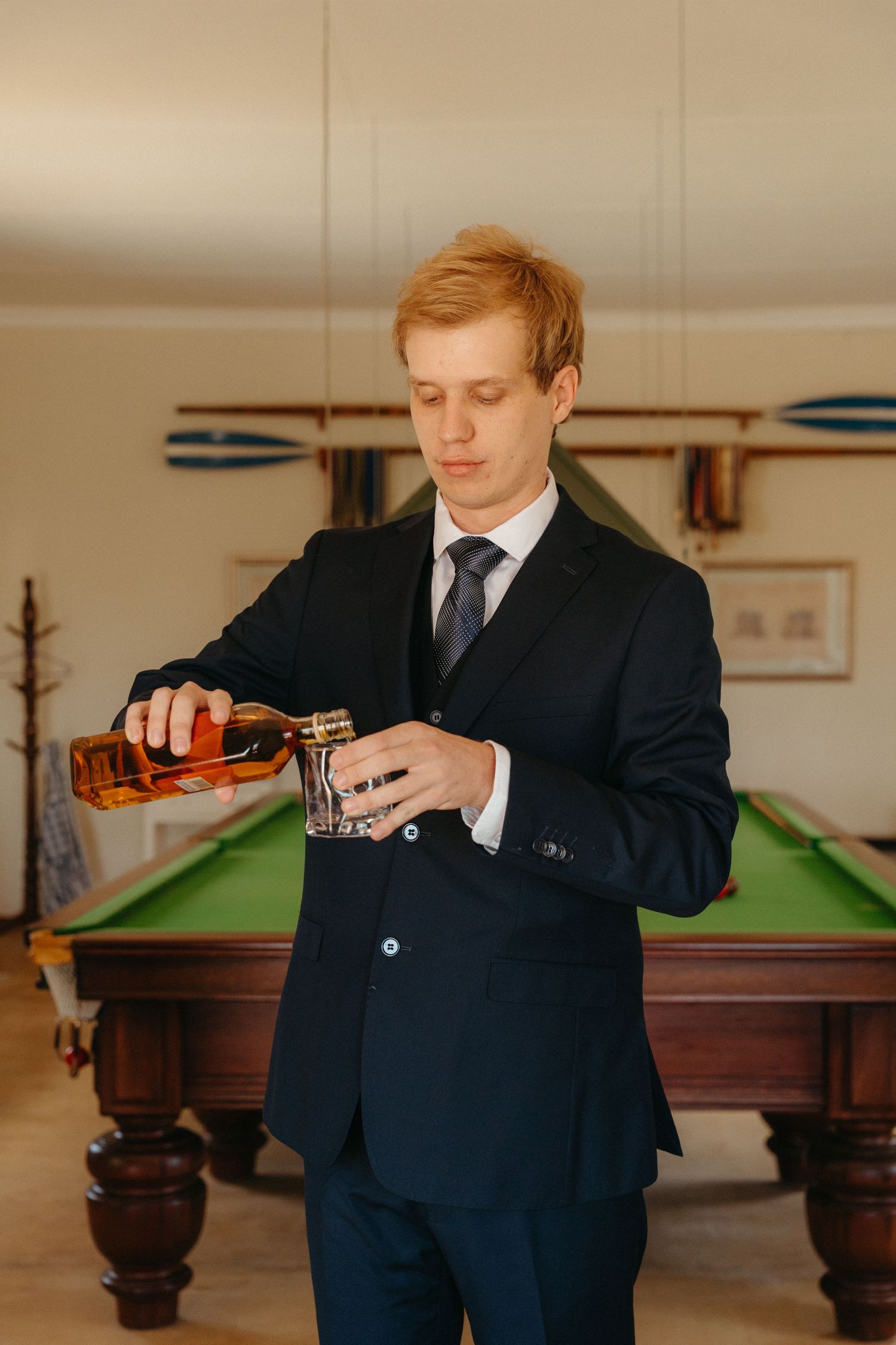 A person in a dark suit pours a drink at a billiards room with a pool table in the background.