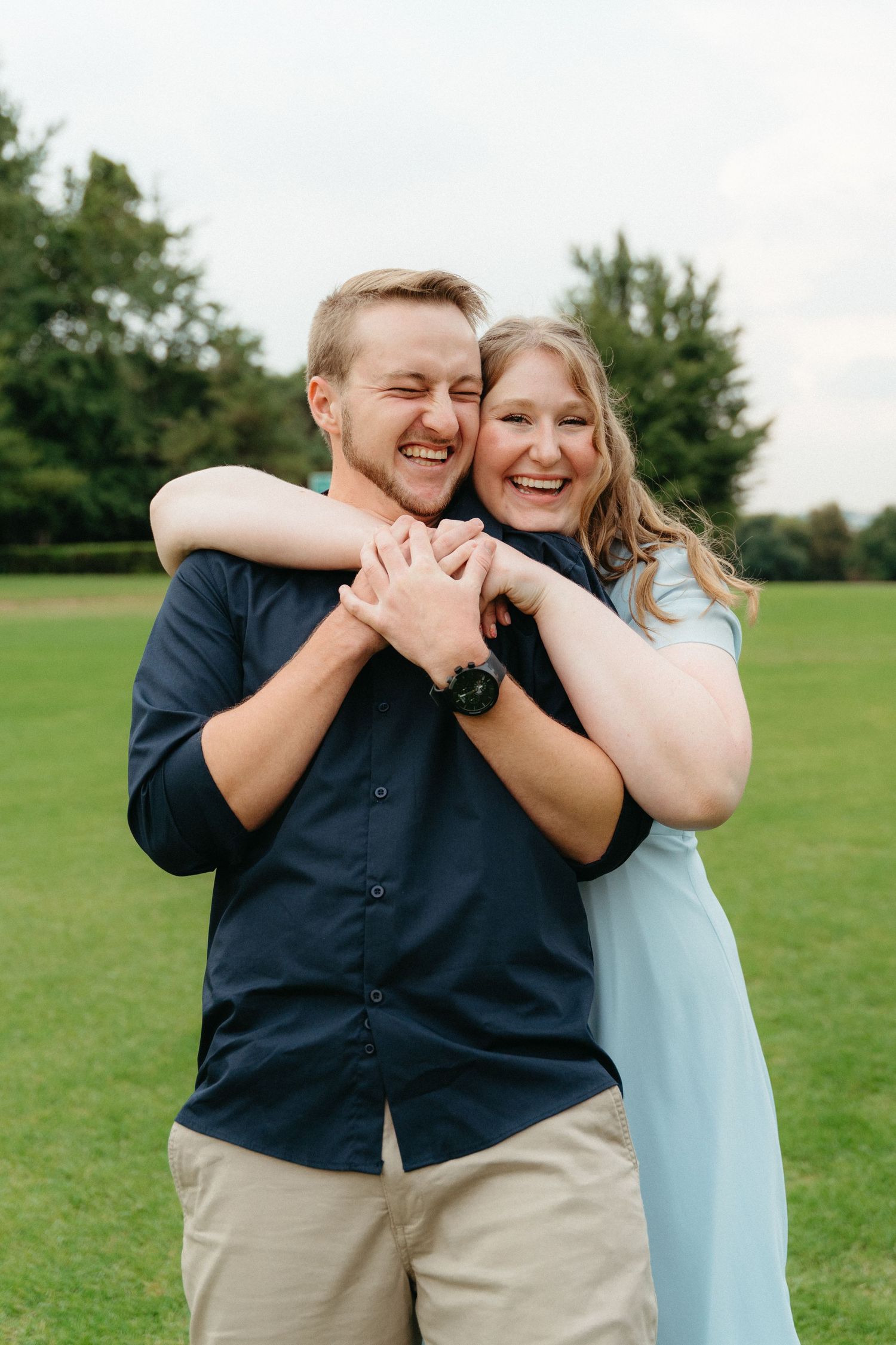 A couple laughs together while embracing on a grassy field on a cloudy day.