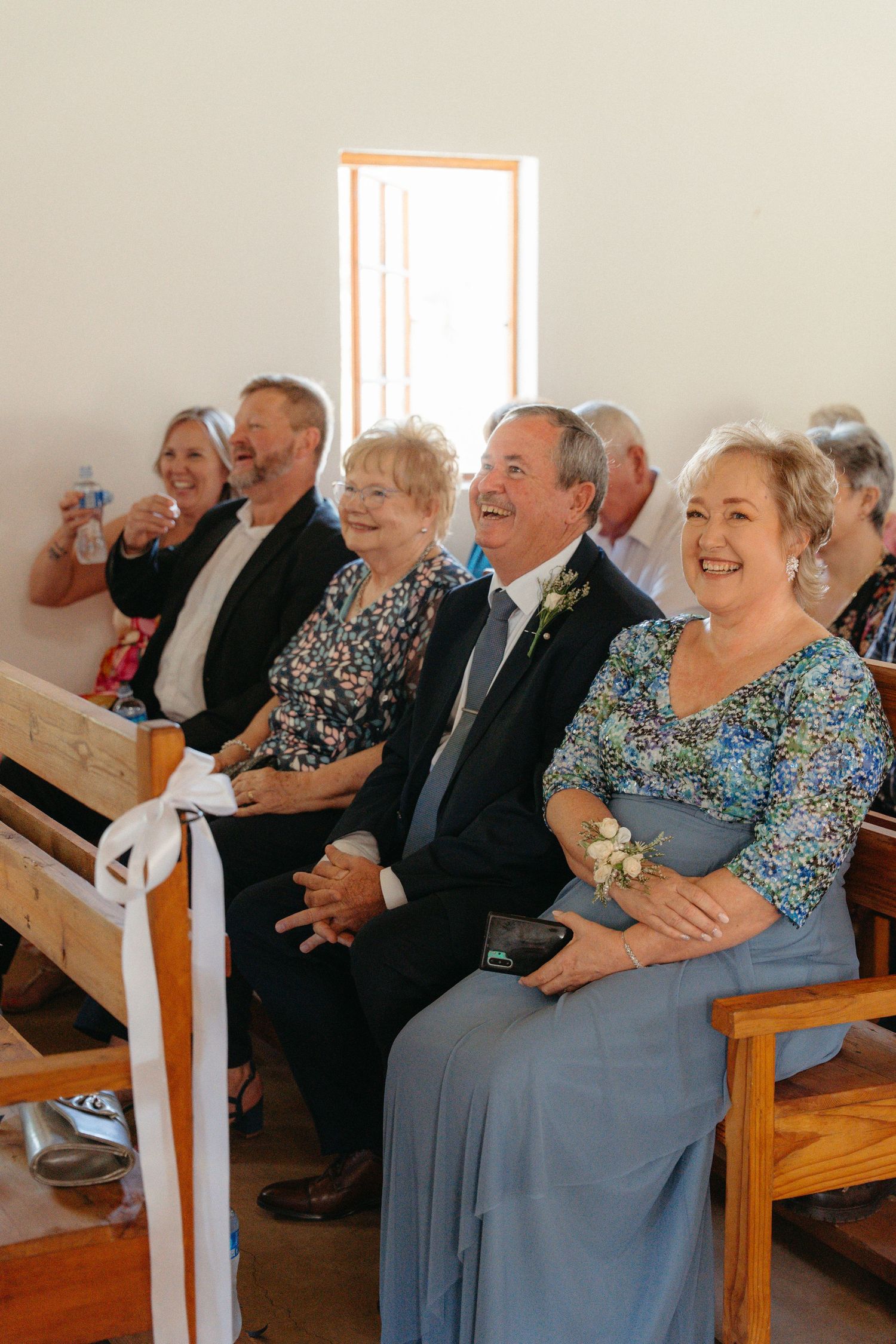 Wedding guests seated in wooden church pews share joyful laughter during the ceremony.