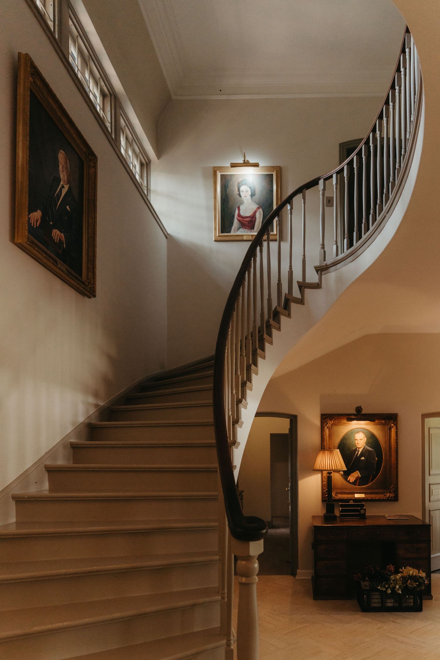 Beechwood Gardens elegant curved staircase with dark wood railing, portrait paintings on walls, and a classic lamp