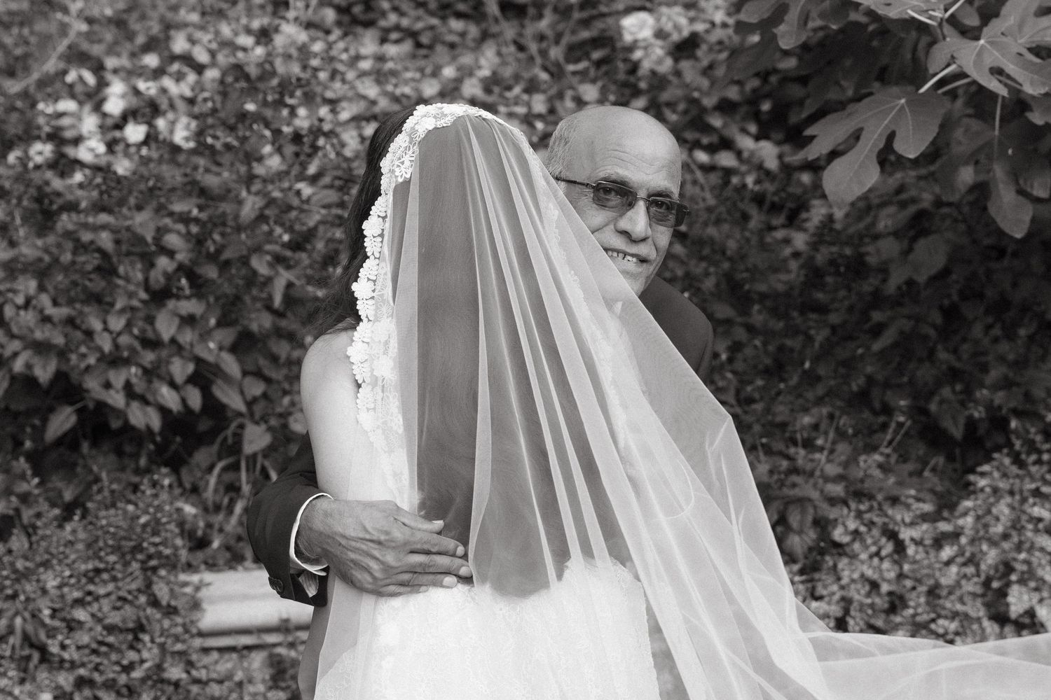 Black and white photo of a bride in a veil embracing an older man in sunglasses outdoors.
