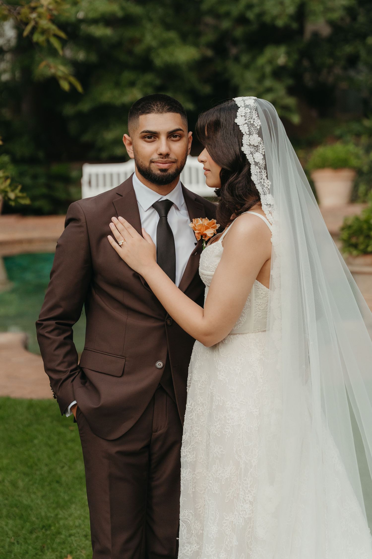 Bride in lace gown and veil stands with groom in brown suit outdoors at garden wedding venue.