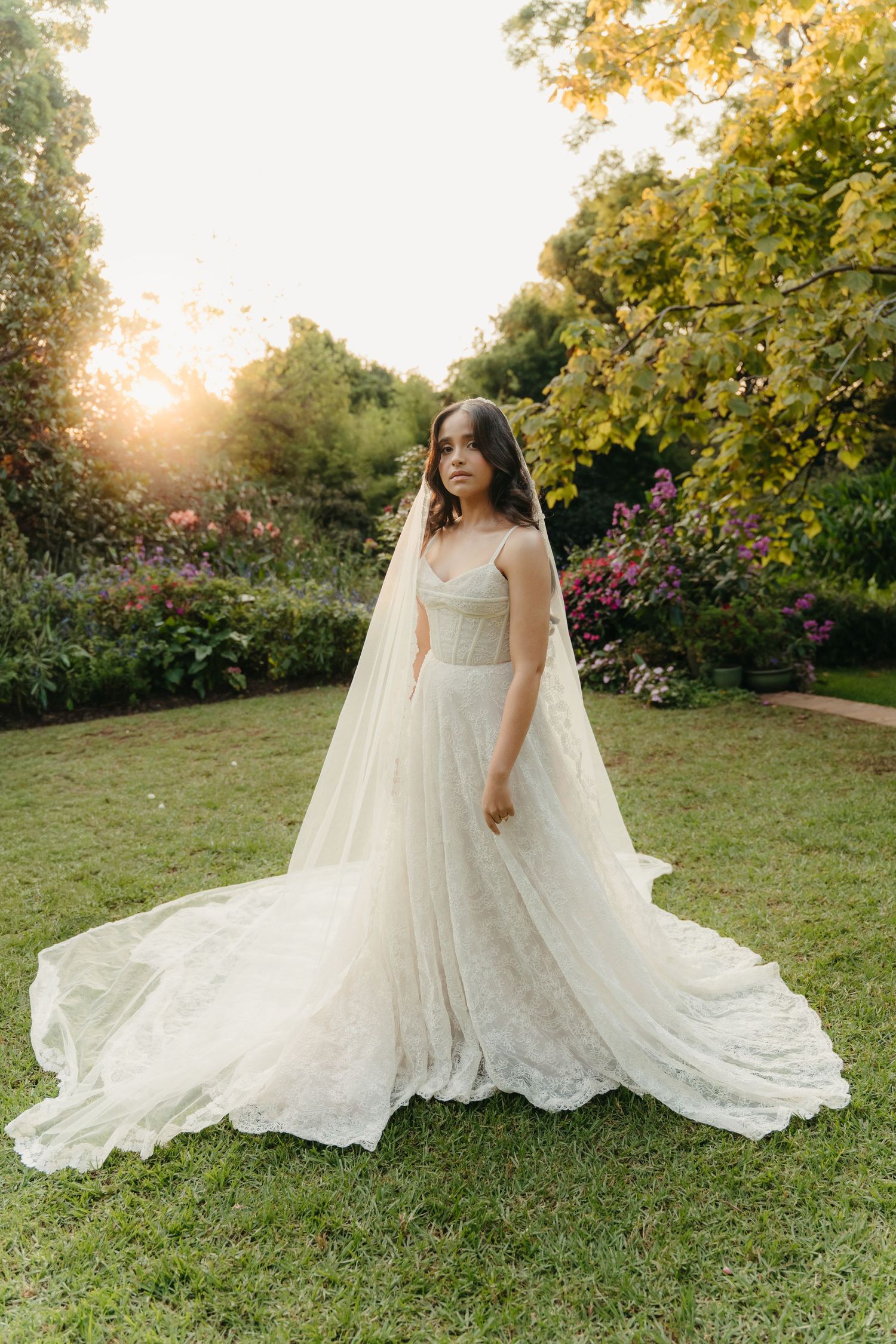 Bride in a flowing white lace gown with a long veil stands in a sunlit garden surrounded by colorful blooms.