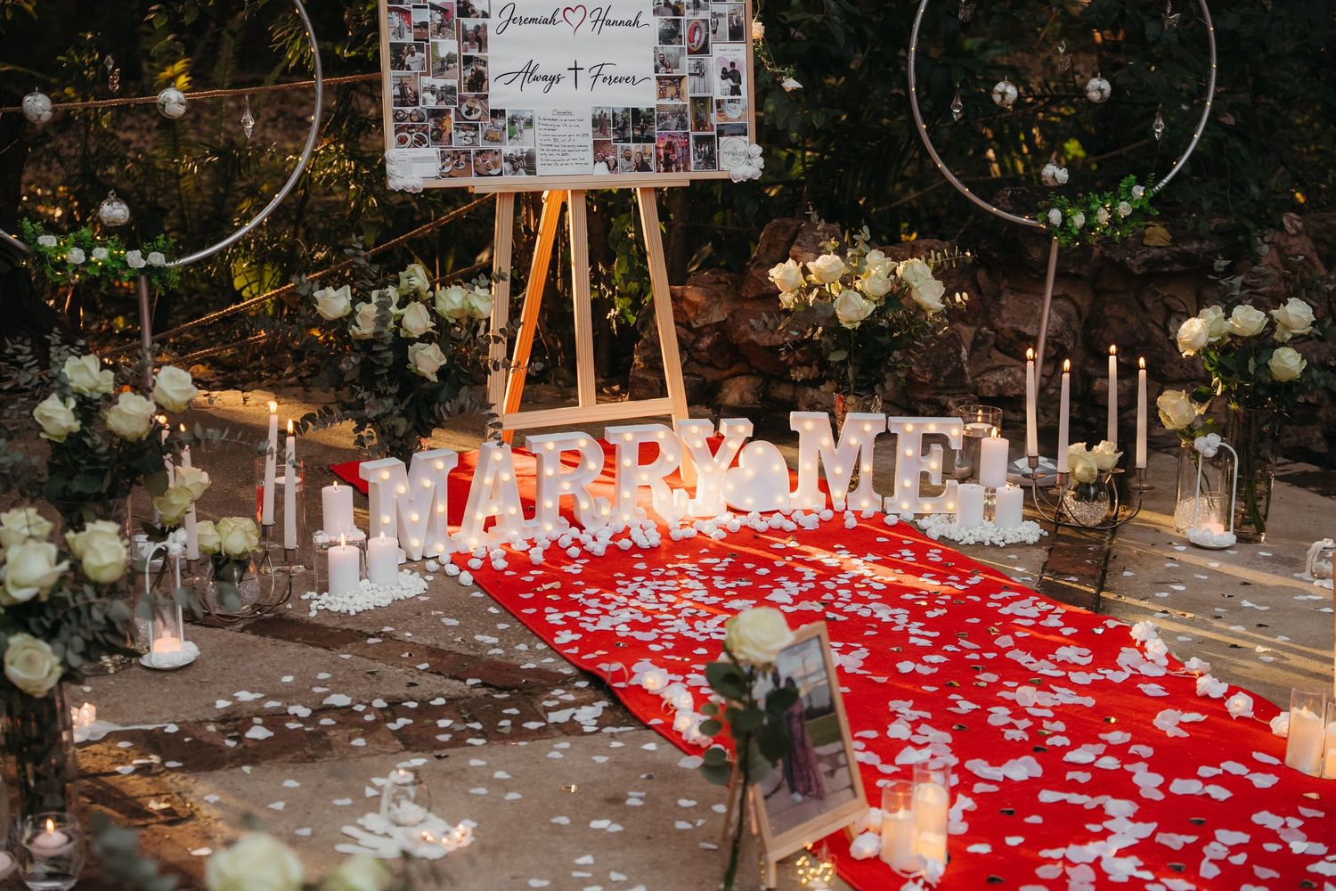 Romantic marriage proposal setup with red rose petal aisle, MARRY ME letters, candles, floral hoops, and a personalized photo display board.