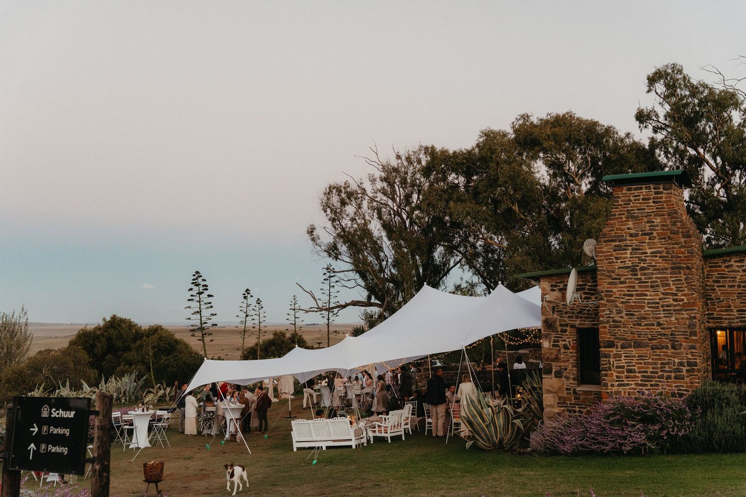 Outdoor wedding reception under white stretch tent beside stone building at dusk with guests mingling on green lawn.