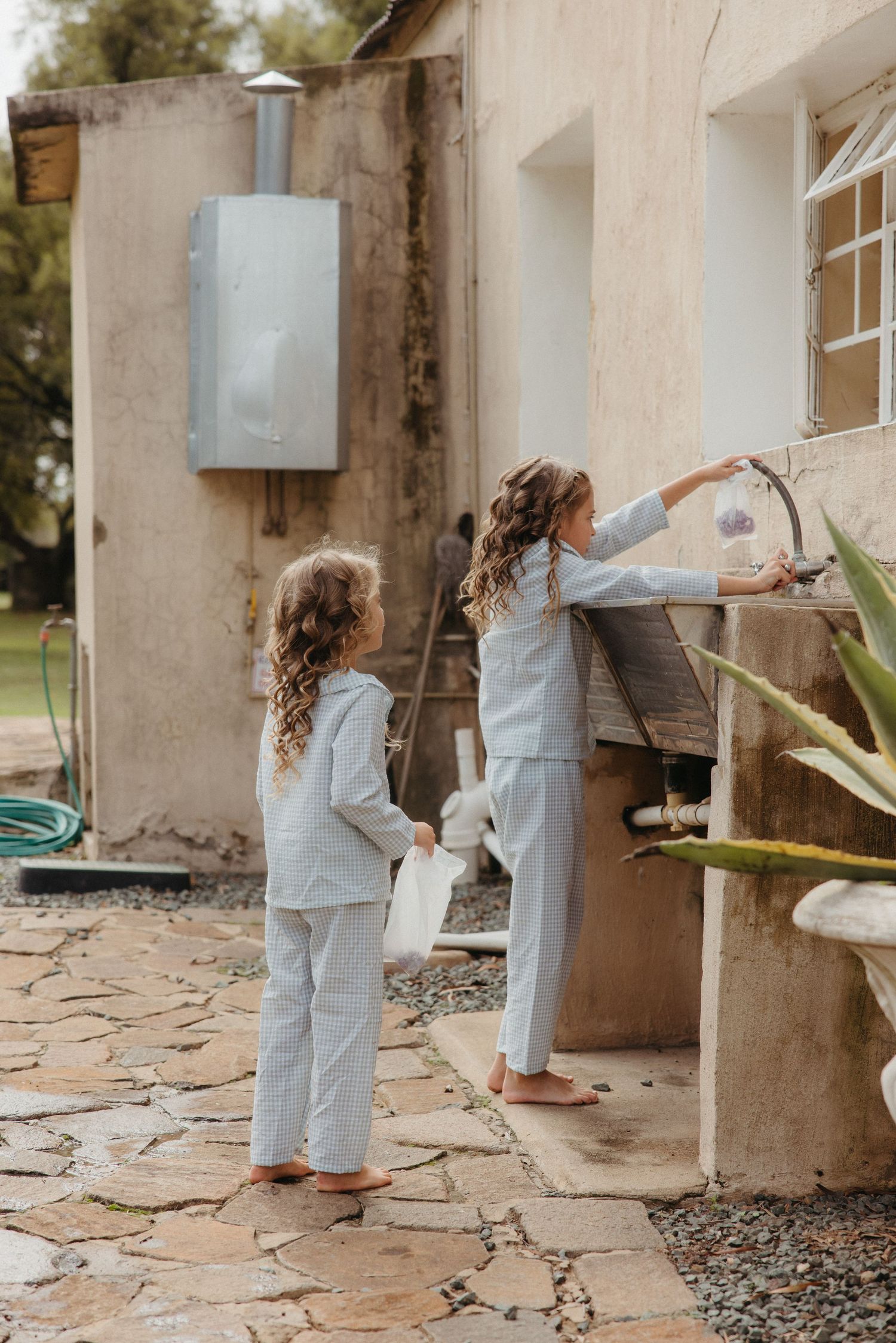 Two girls in matching light blue pajamas washing hands at an outdoor sink beside a rustic building.