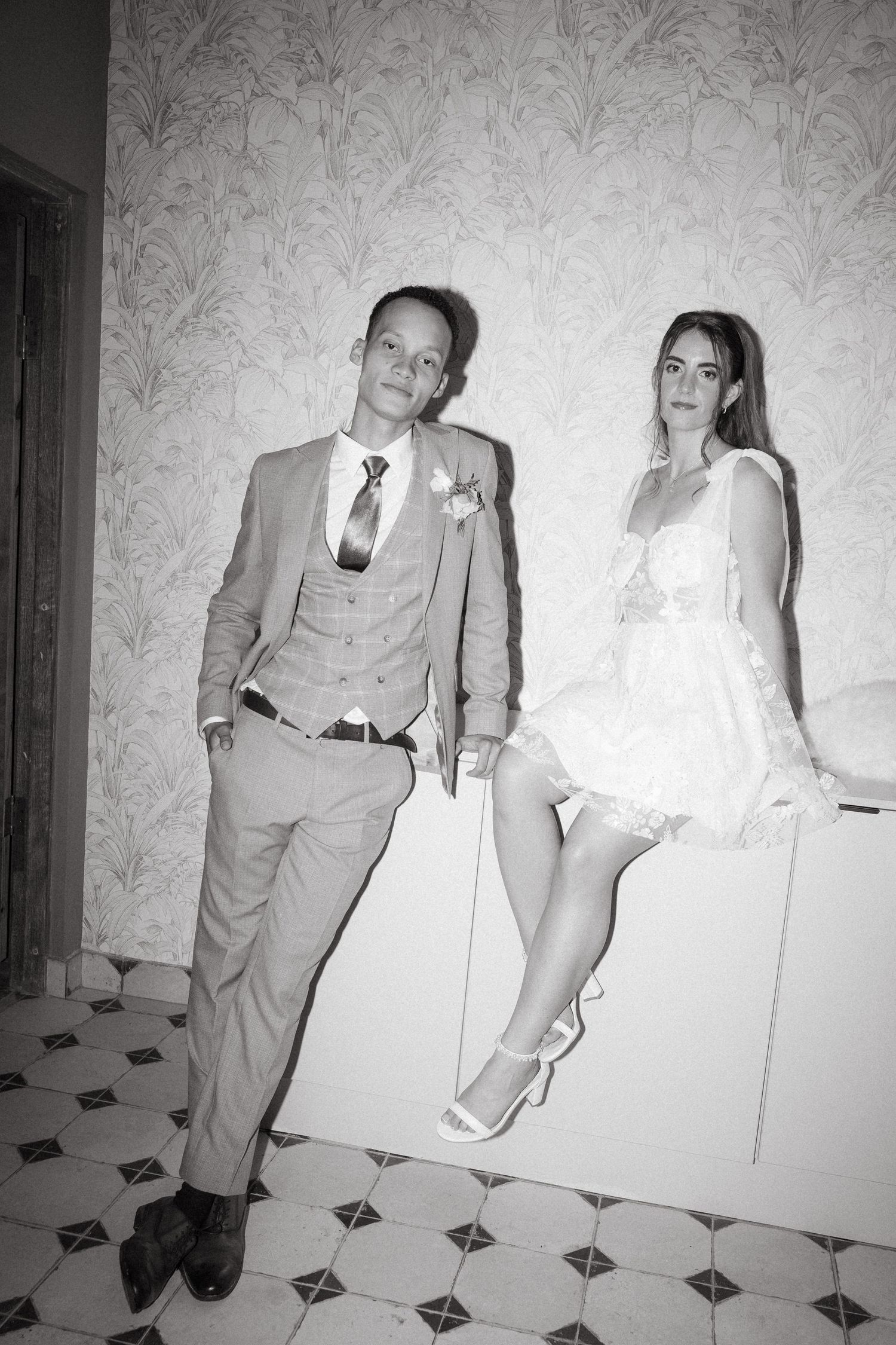 Black and white photo of a stylish couple posing, groom in suit with tie, bride in white lace dress, elegant interior.