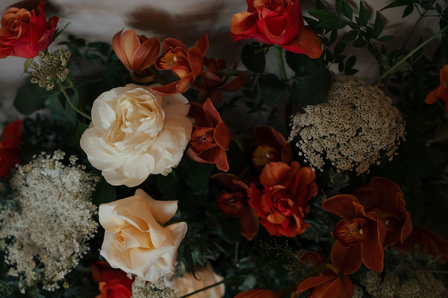 Wedding floral arrangement with cream roses, rust orange tulips, white Queen Anne's lace and dark green foliage in warm autumn tones.