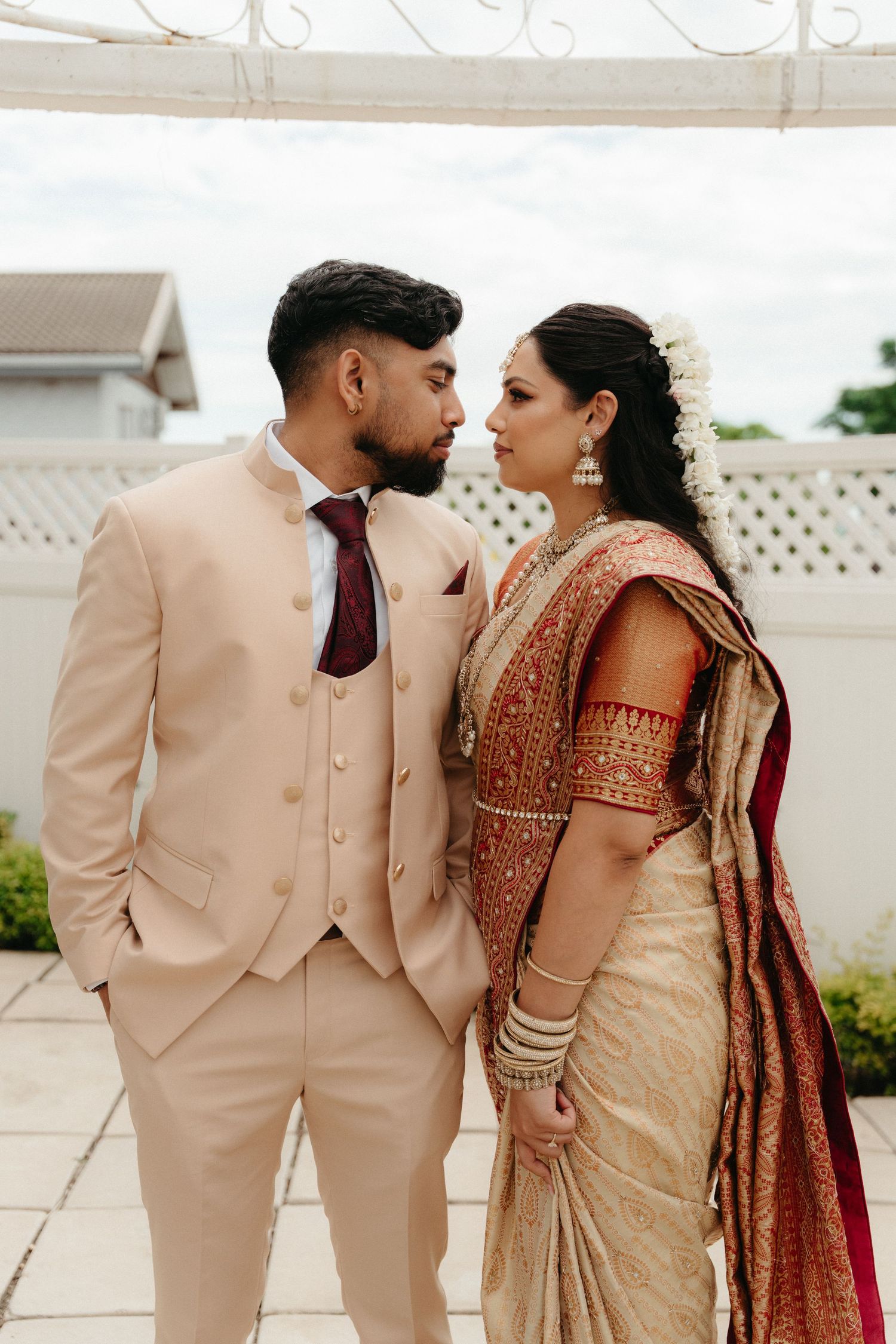 A couple in wedding attire featuring a beige suit and traditional Indian bridal saree pose together outdoors.