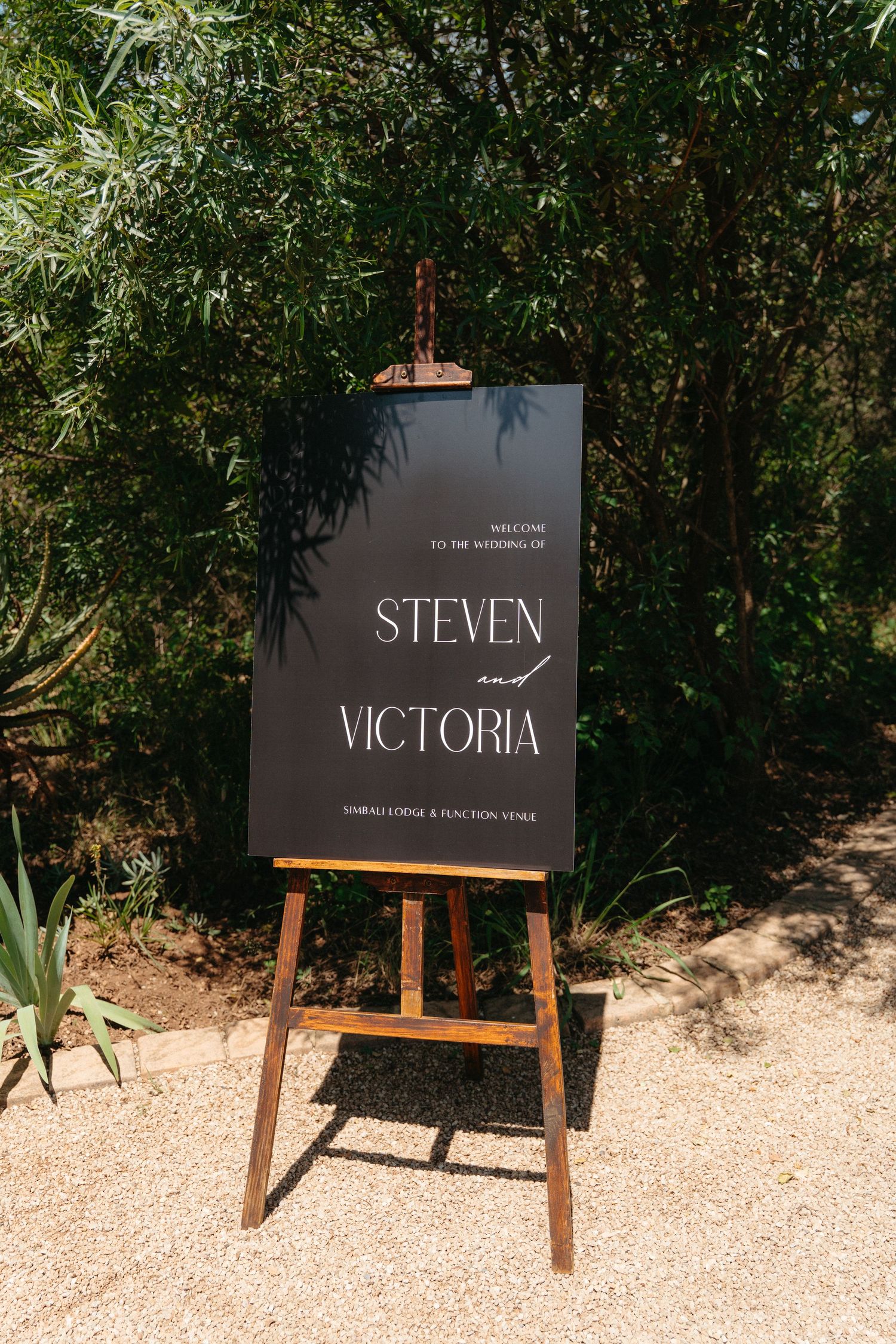 A black wedding welcome sign for Steven and Victoria displayed on a wooden easel in a garden setting.