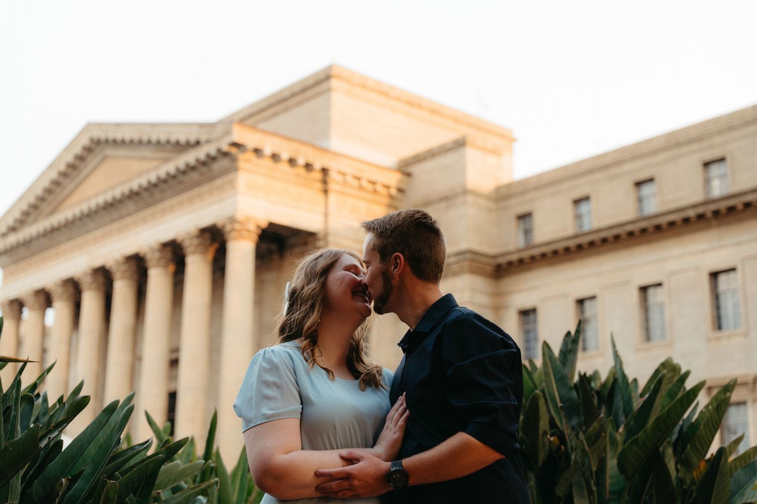 A couple shares a romantic kiss on a bench surrounded by tropical plants with a grand building facade in the background.