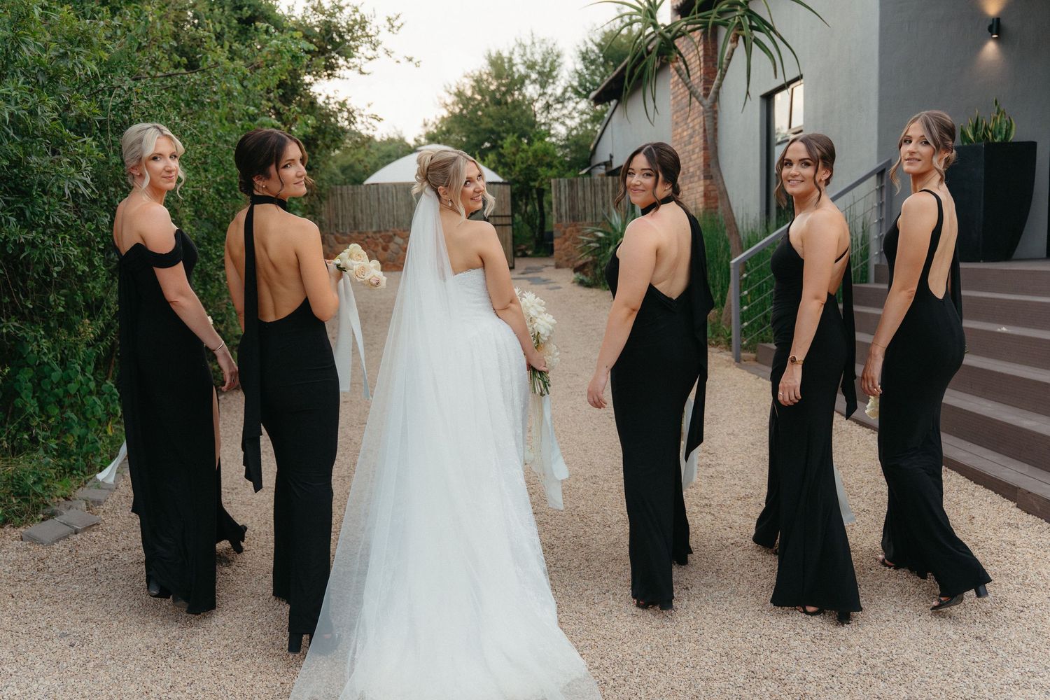Wedding party wearing black dresses and white gown walk together along outdoor path at sunset.