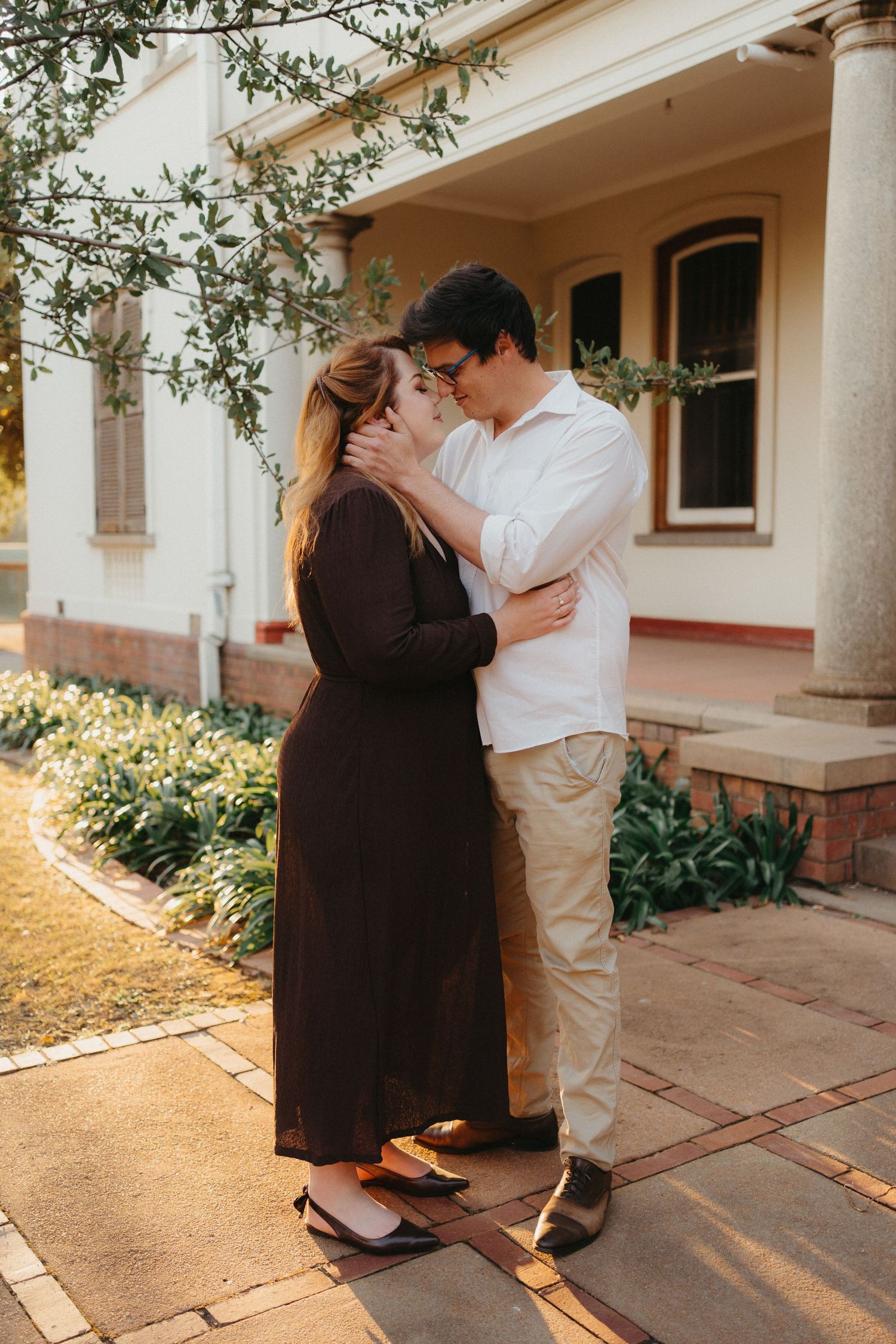 Couple sharing a tender embrace and kiss in a sunlit garden beside a heritage building in Pretoria