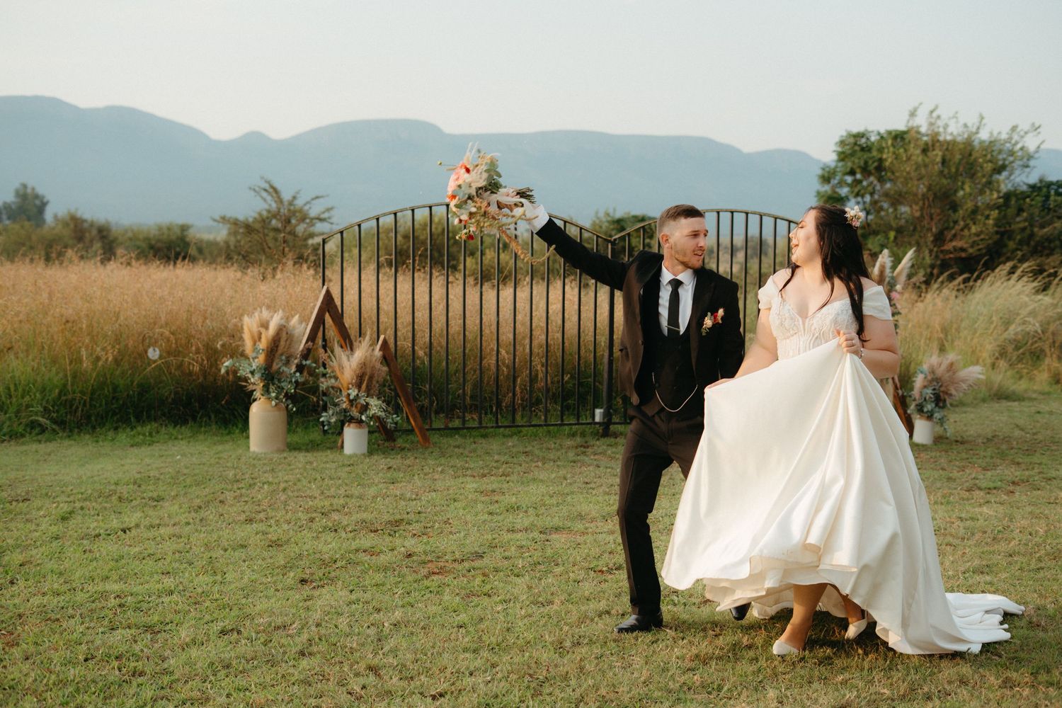 Groom lifts bride in white gown outdoors at Stable View Venue with Hekpoort mountains and golden Gauteng fields in the background.