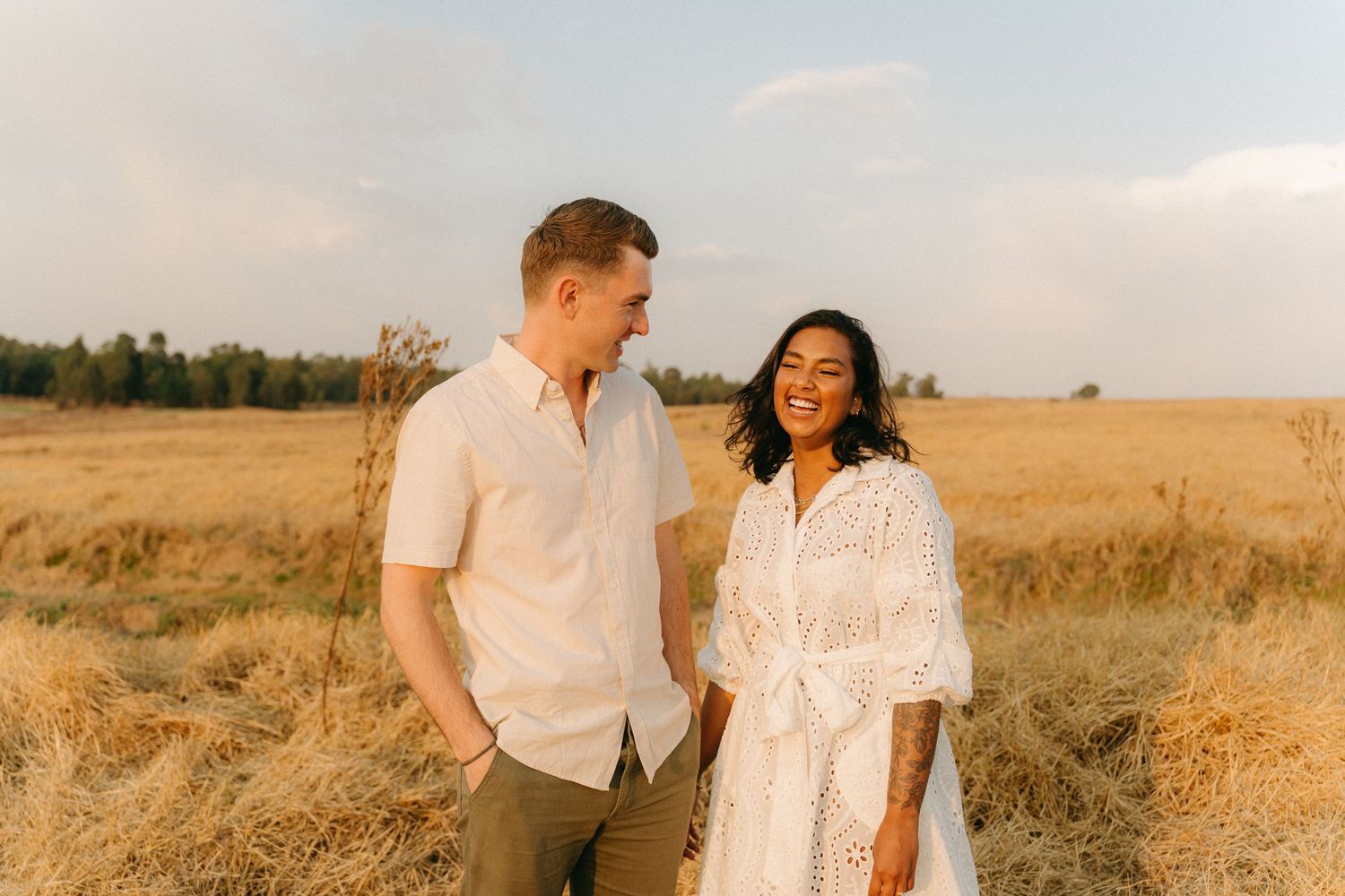 Couple in white outfits laughing together in a sunlit meadow during golden hour.