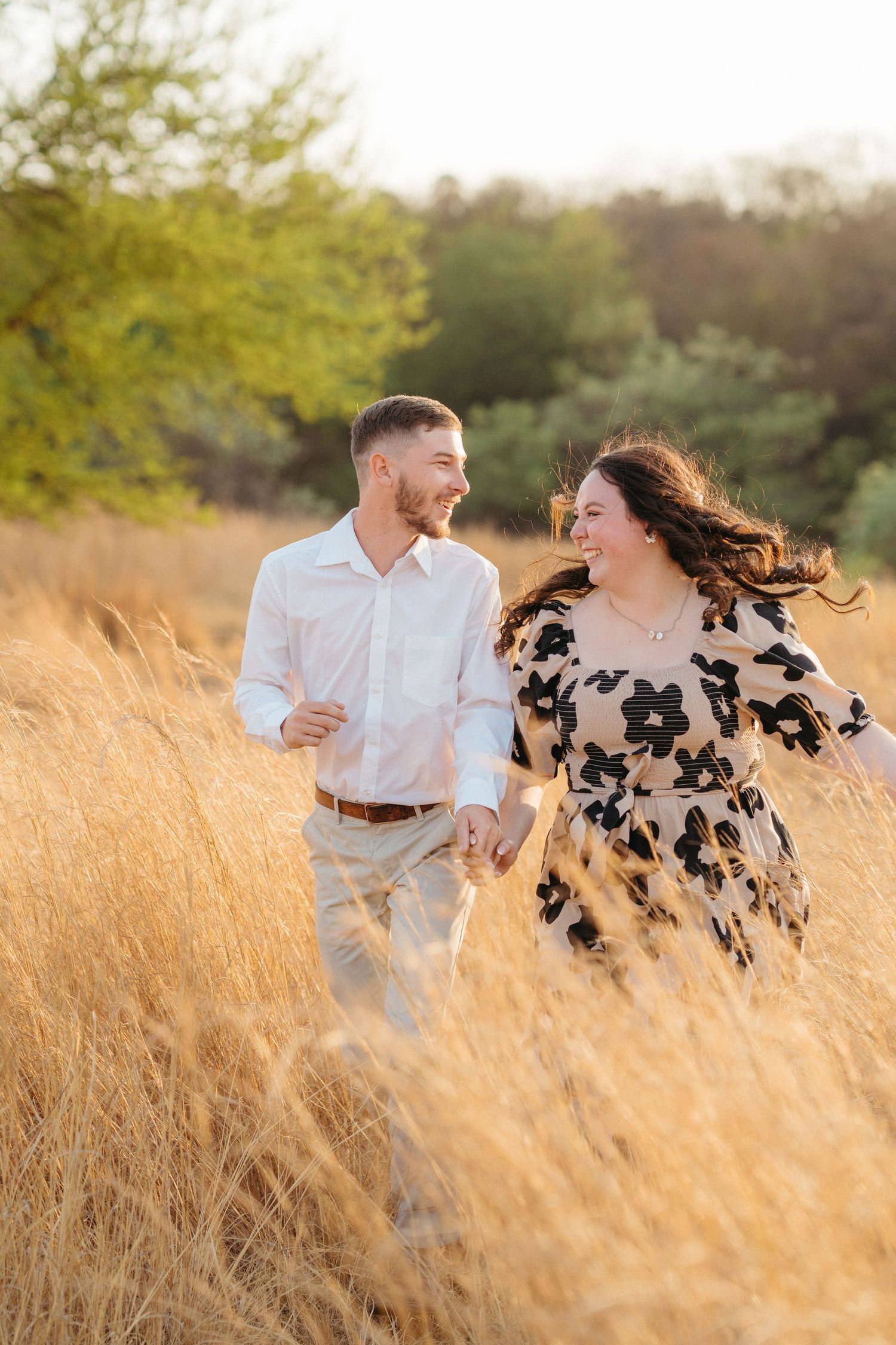 Couple running and laughing together through tall golden grass at sunset in a romantic outdoor setting.
