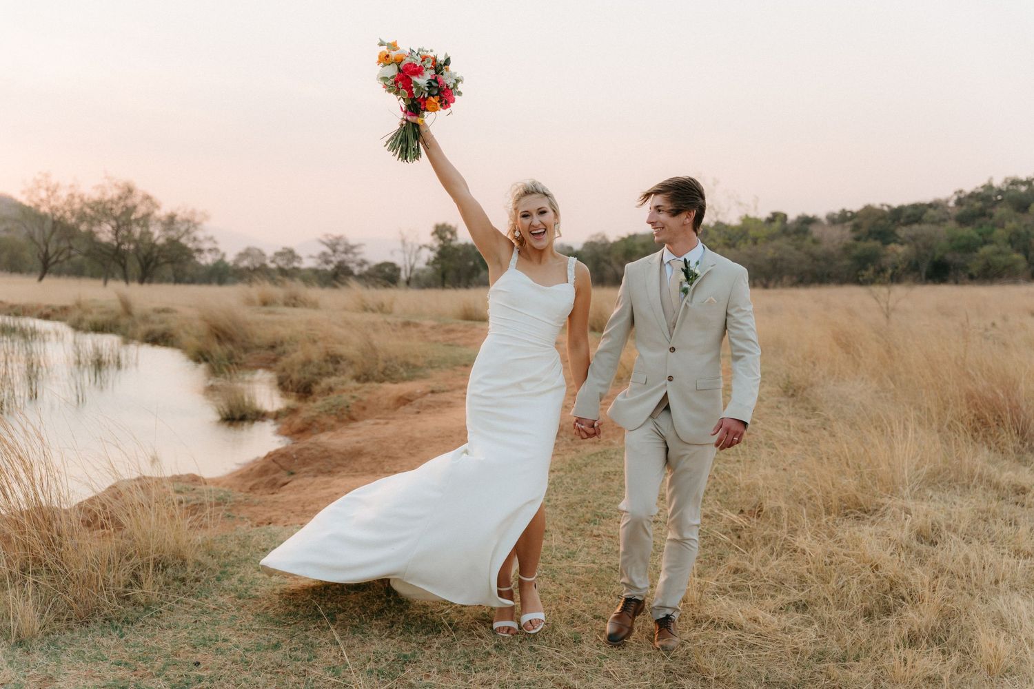 Couple in wedding attire dancing joyfully in a rustic outdoor setting during golden hour.