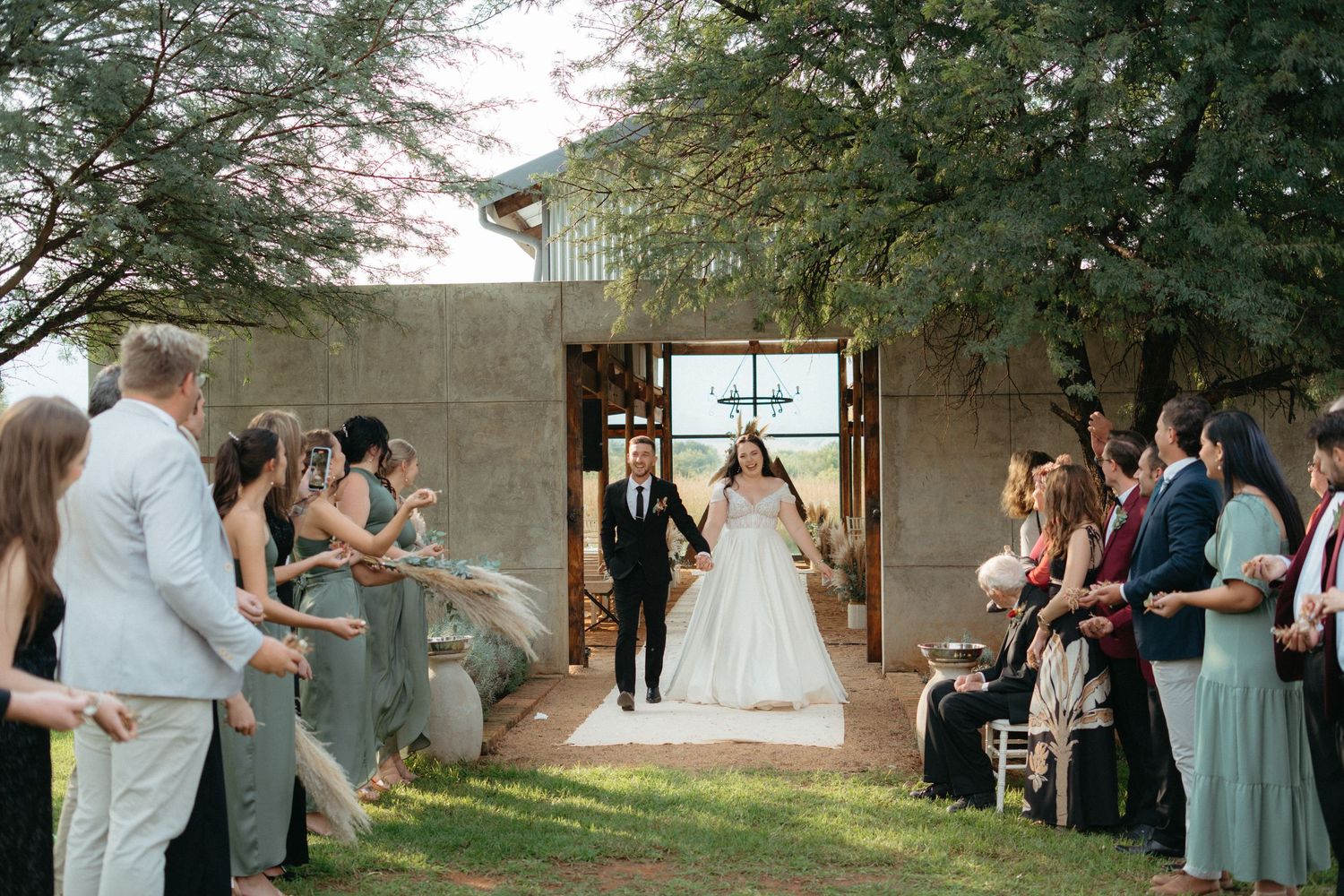 Wedding ceremony underway at modern outdoor venue with guests watching couple exchange vows under rustic archway.