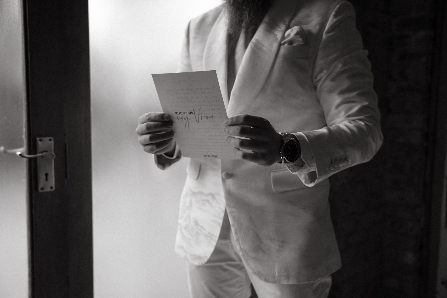 A groom in a white suit holds papers while standing in dramatic black and white lighting.