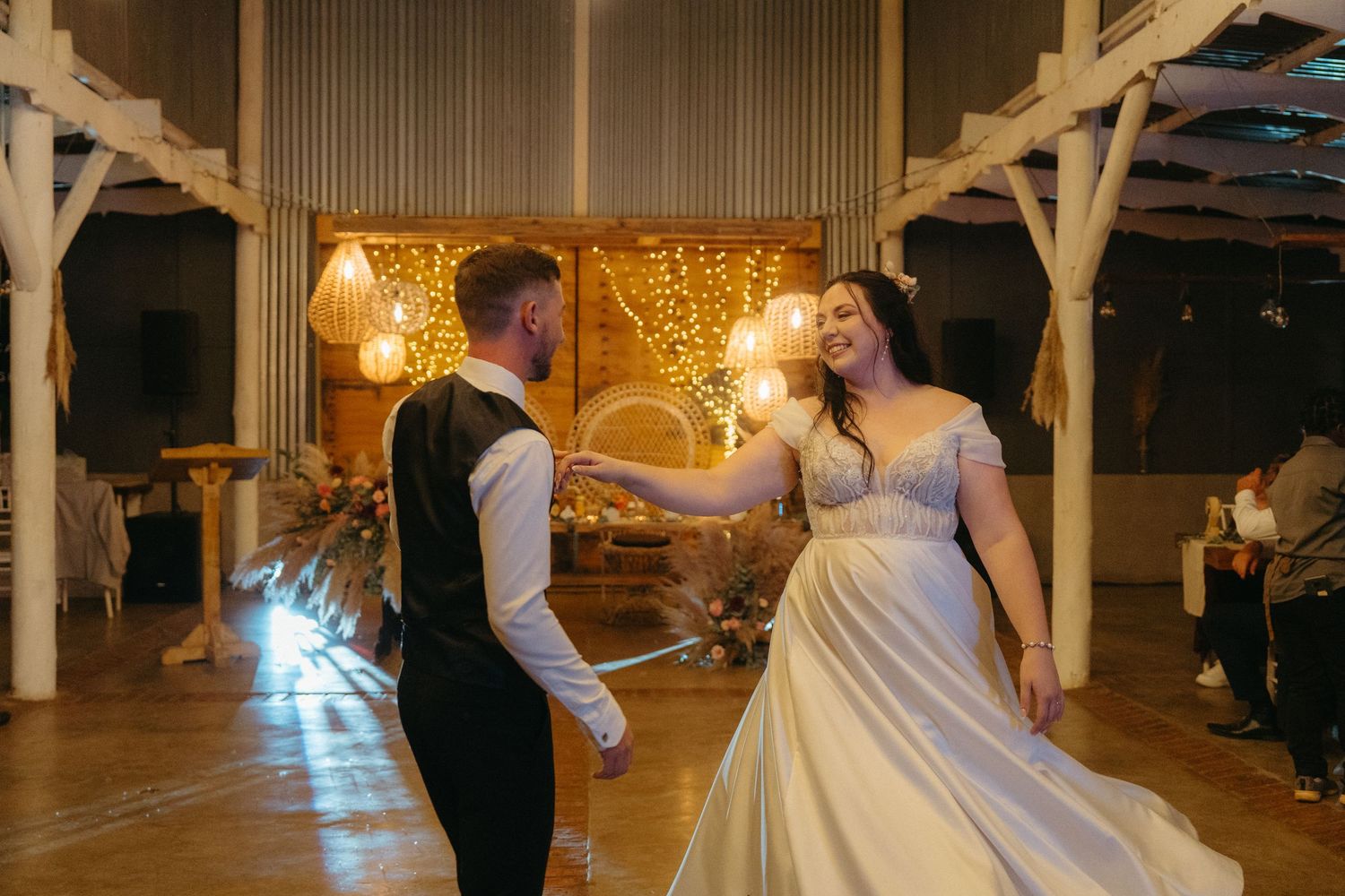 A couple dances in an elegantly lit barn venue with twinkling lights and wooden beams.