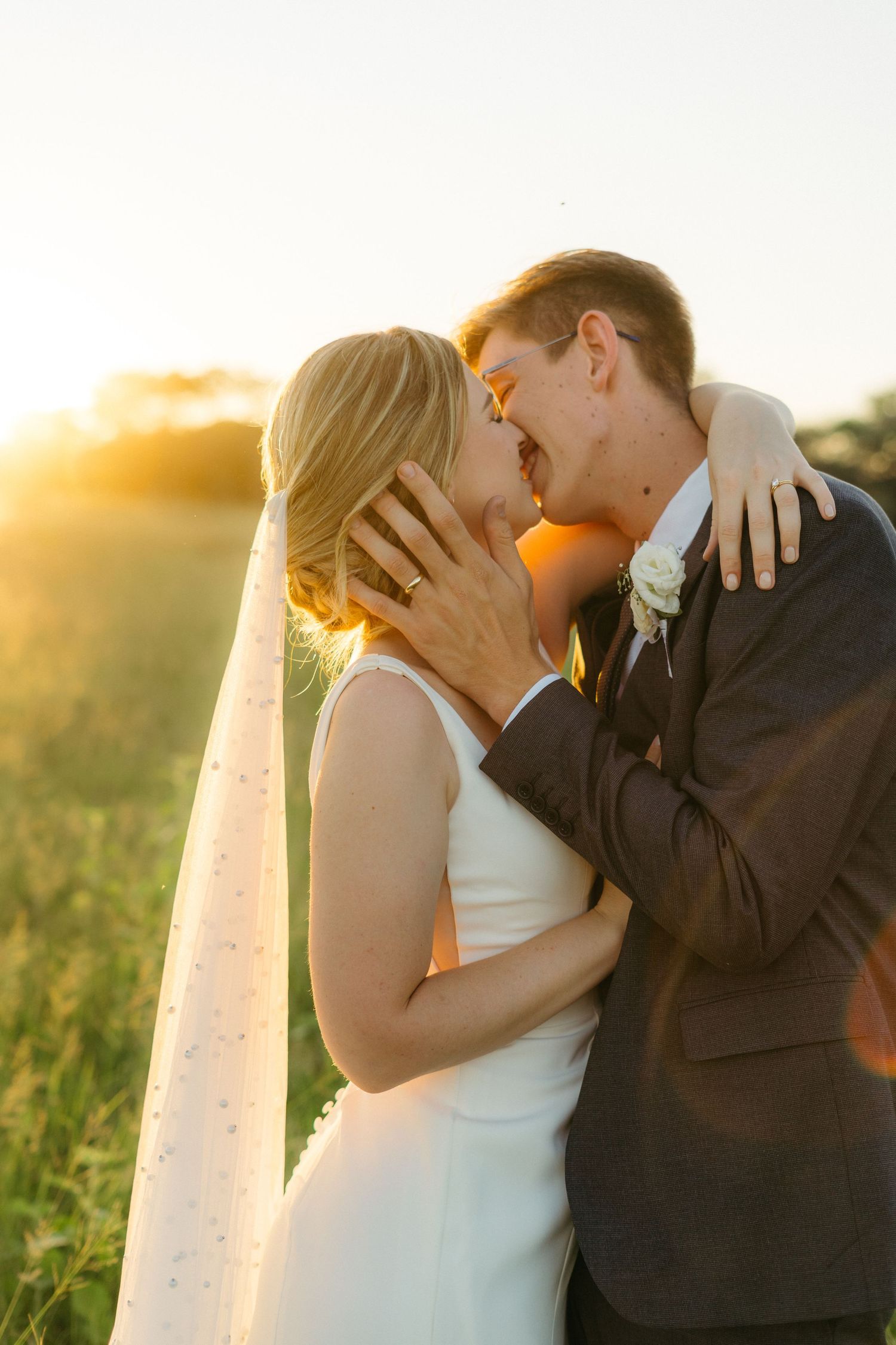 Close-up of newlyweds sharing an intimate kiss during golden hour with long veil and brown suit in a field.