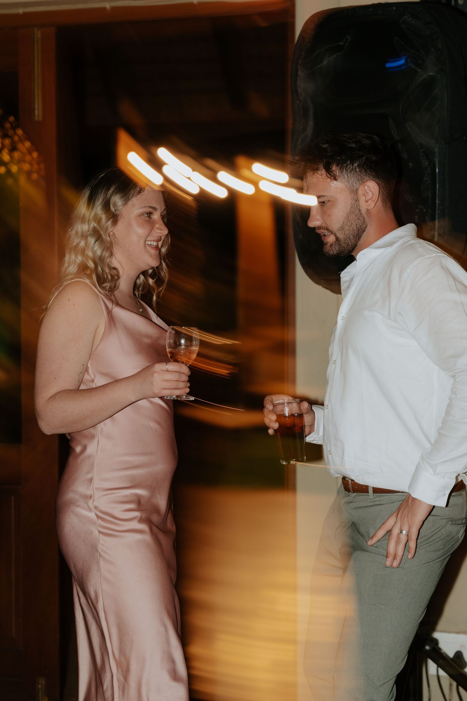 A couple in formal attire share a candid moment at a party, with blurred lights in the background.