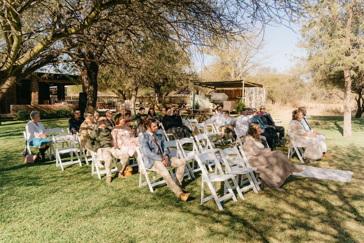 Wedding guests seated in white chairs under trees during an outdoor ceremony.