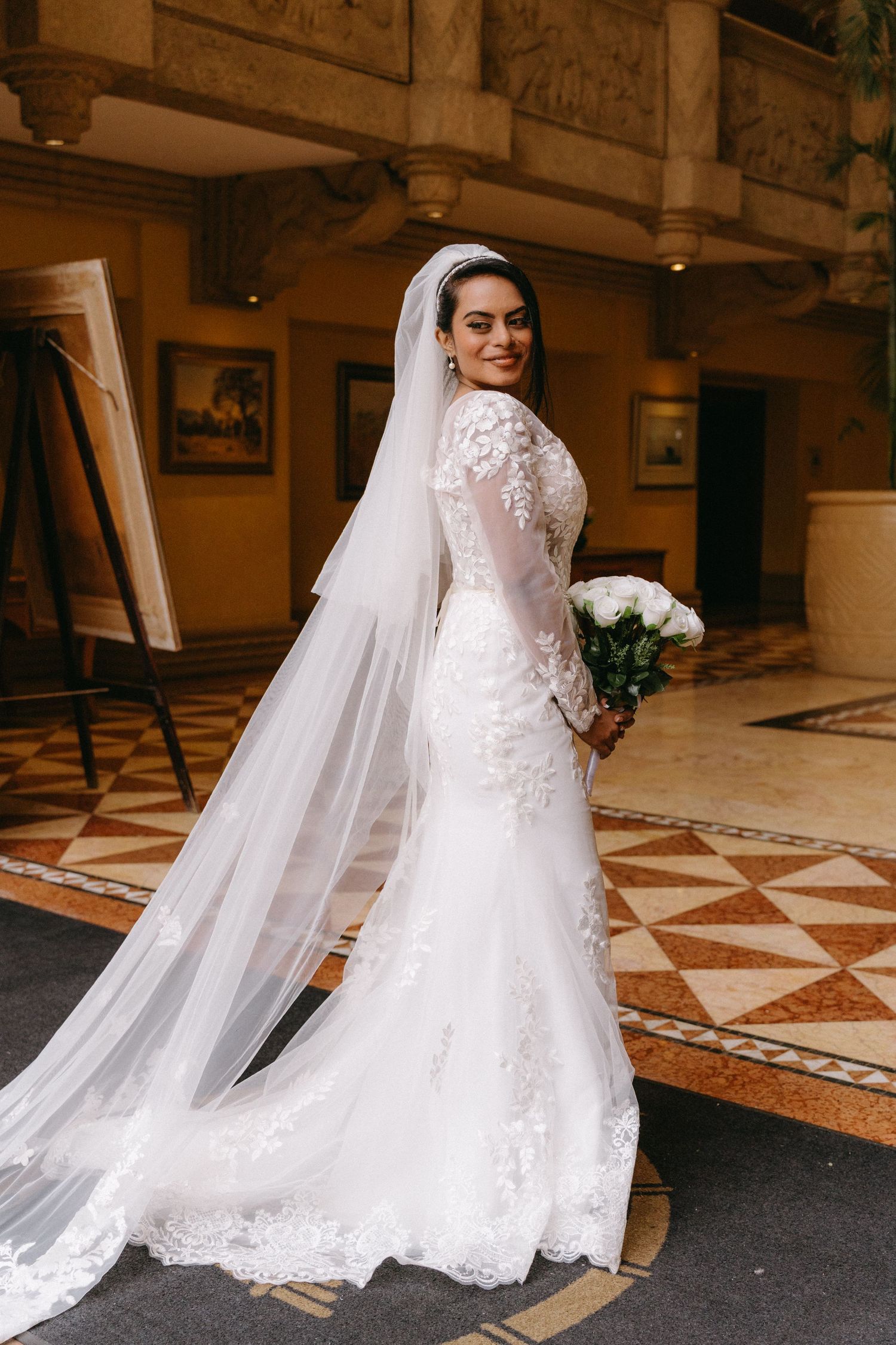 A bride in a stunning lace-detailed mermaid wedding dress and long veil poses elegantly in an ornate hallway.